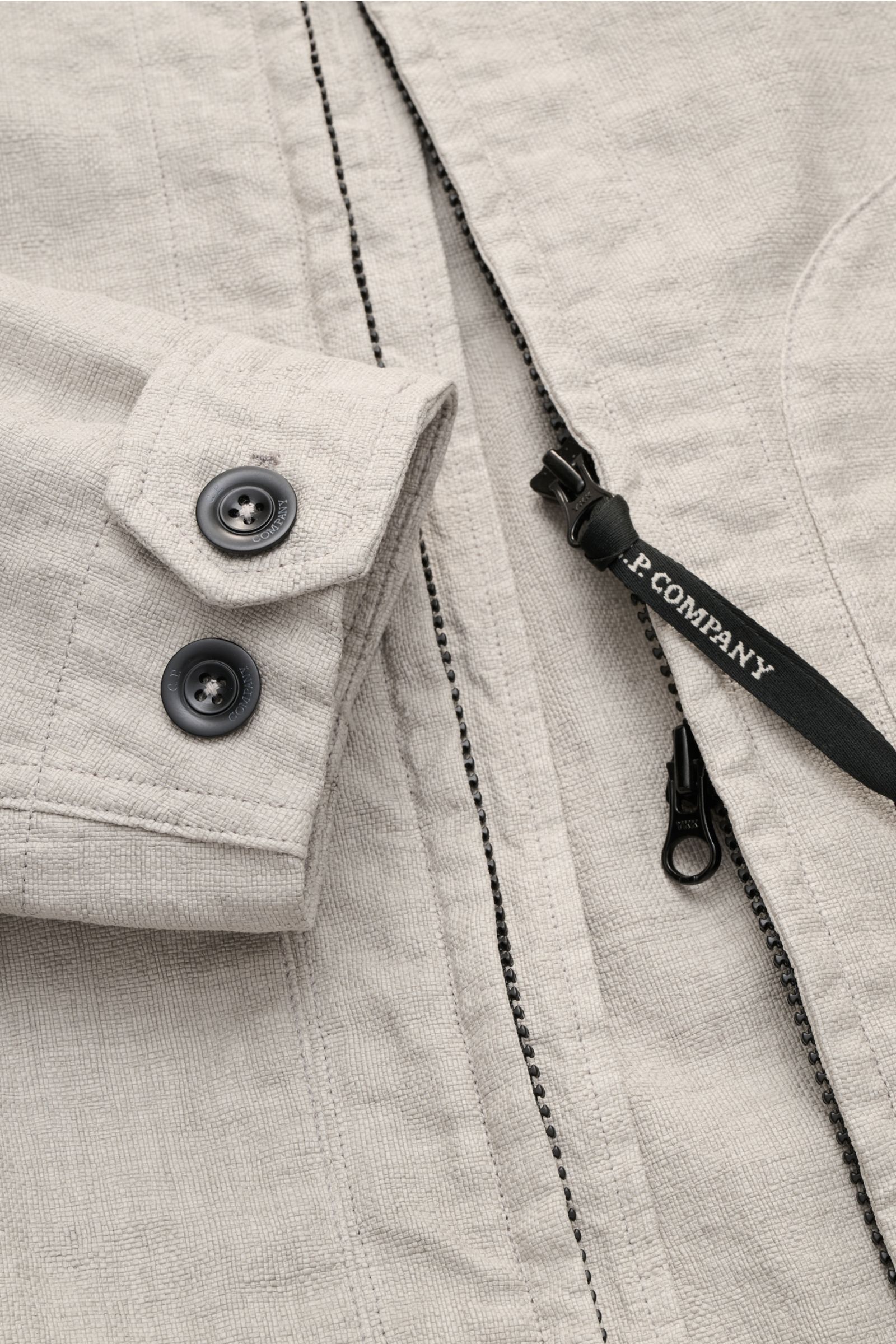 Close-up front view of C.P. Company Jacket 'Goggle Jacket' grey showing textured high-quality microfibre, buttons on cuff, and robust two-way zip with branded pull. Signature design from C.P. COMPANY: The 'Goggle Jacket' makes a futuristic statement with 