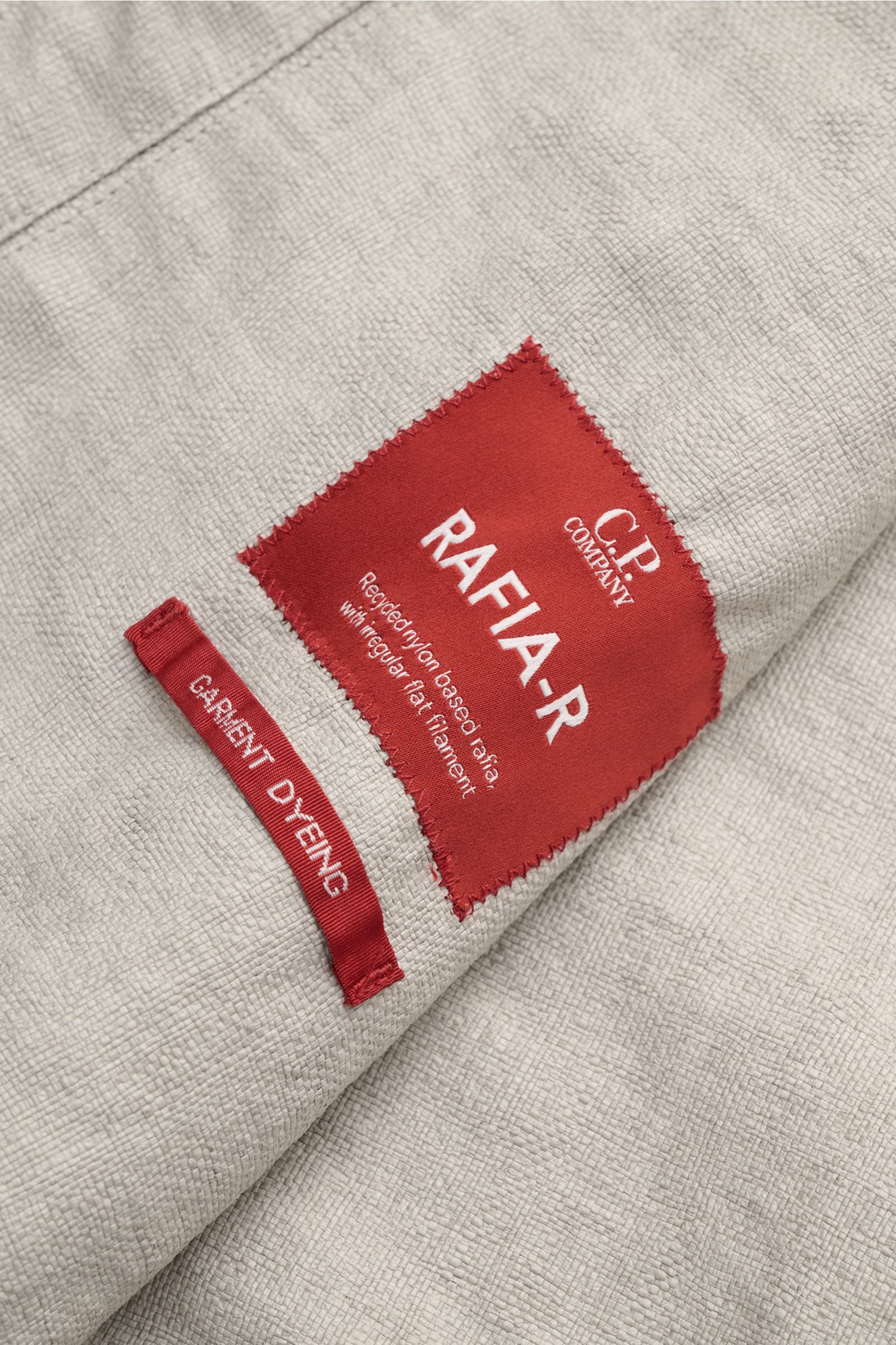 Close-up of the textured fabric and red label of the C.P. Company Jacket 'Goggle Jacket' grey, photographed from above.

Signature design from C.P. COMPANY: The 'Goggle Jacket' makes a futuristic statement with its goggle lenses integrated into the hoo