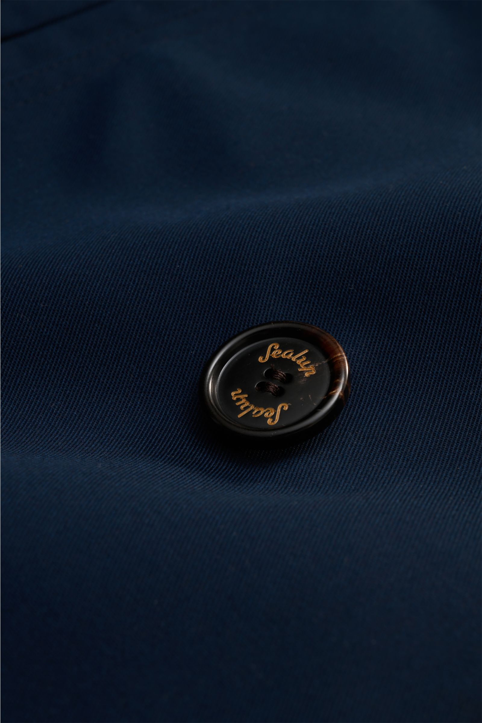 Close-up front view of the Sealup Pea coat 'Amalfi' navy showing a dark button with gold 'Sealup' logo on fine twill fabric. Get the look of a Hanseatic merchant with the elegant pea coat 'Amalfi' by traditional label SEALUP: made of lightweight twill in 