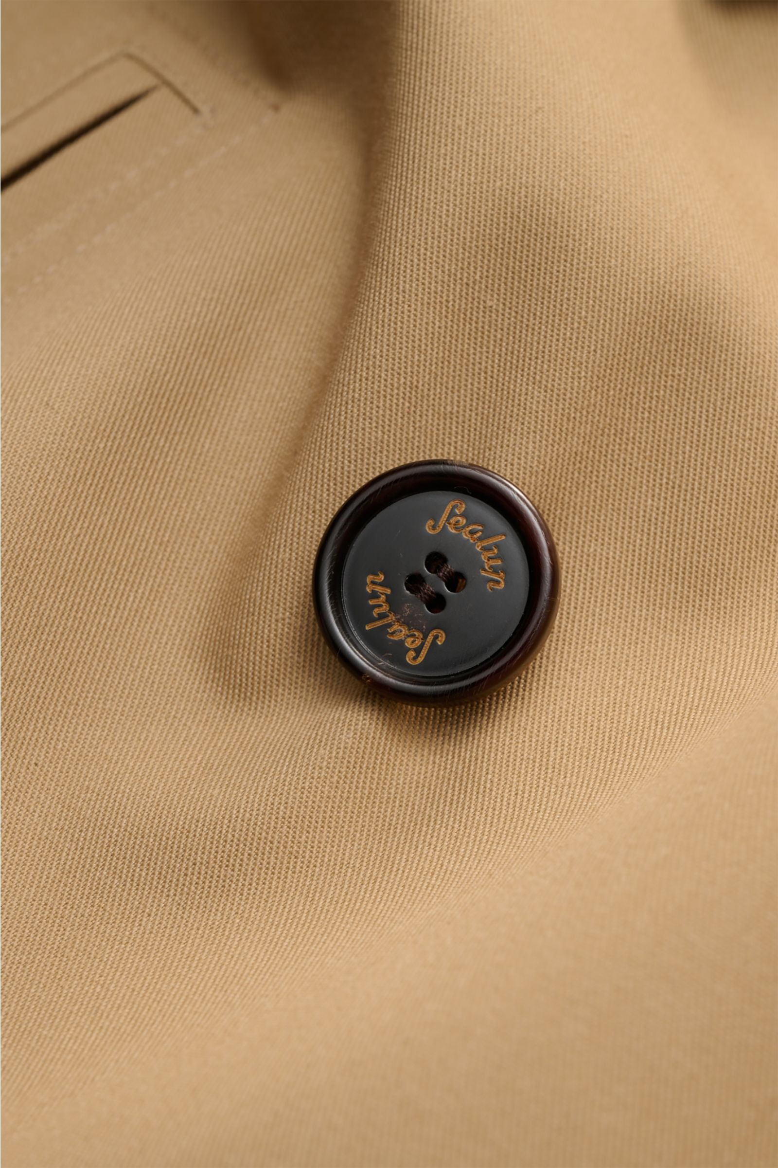 Close-up front detail of the Sealup Pea coat 'Amalfi' beige, showing a dark button with logo on fine twill fabric from a slightly angled perspective. Get the look of a Hanseatic merchant with the elegant pea coat 'Amalfi' by traditional label SEALUP: made