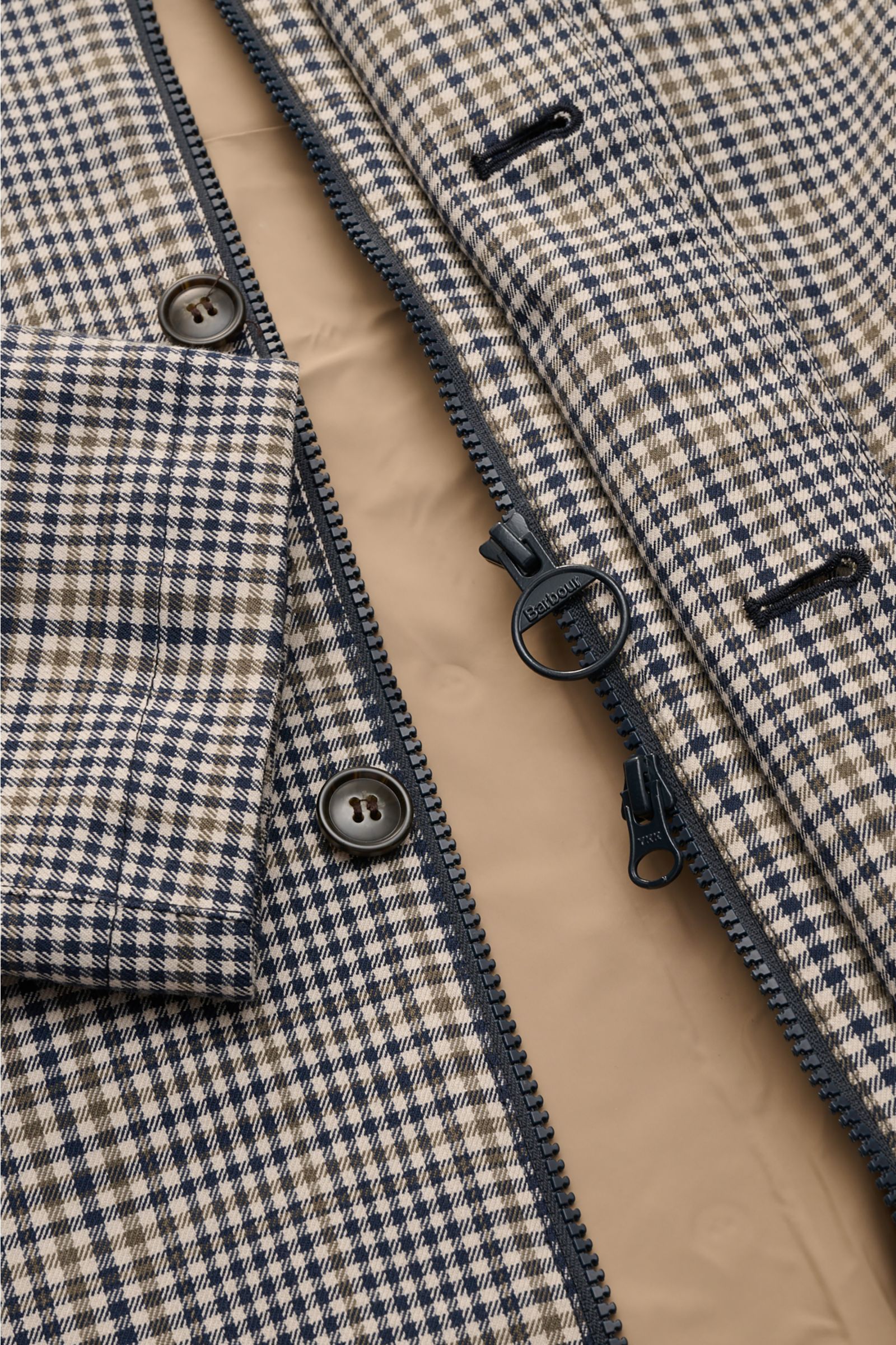 Close-up image from above showing the navy and olive checked pattern of the Barbour Rain coat 'Check Rokig' navy/olive checked, highlighting the two-way zip with a circular Barbour-branded pull, buttoned storm flap, and part of the cuff. Be ready for chan