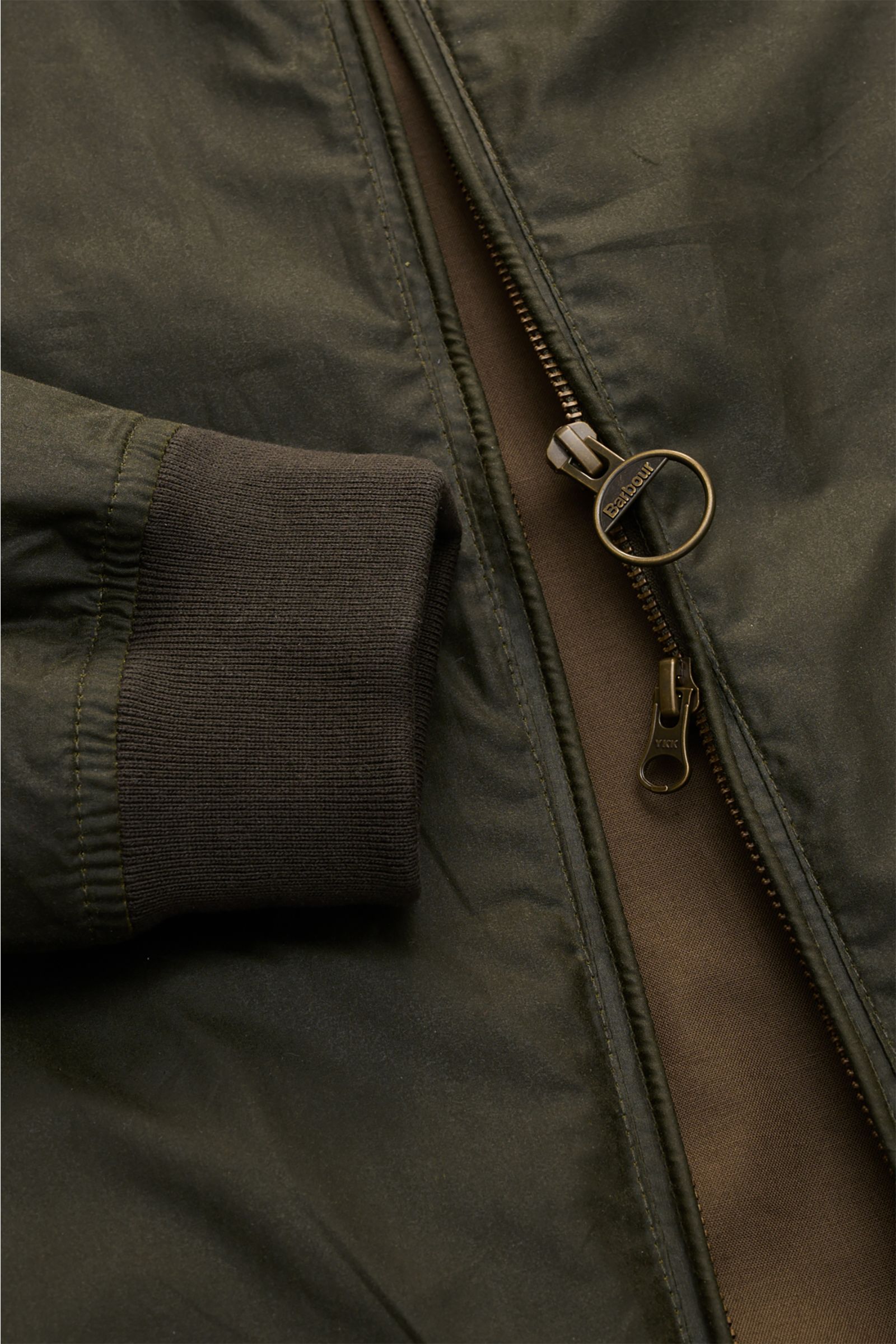Close-up front view of the Barbour Waxed blouson 'Royston' dark olive, showing the partially open two-way zip with a branded ring pull and elasticated ribbed cuff, highlighting the waxed cotton fabric and slim fit design. Classic blouson by traditional