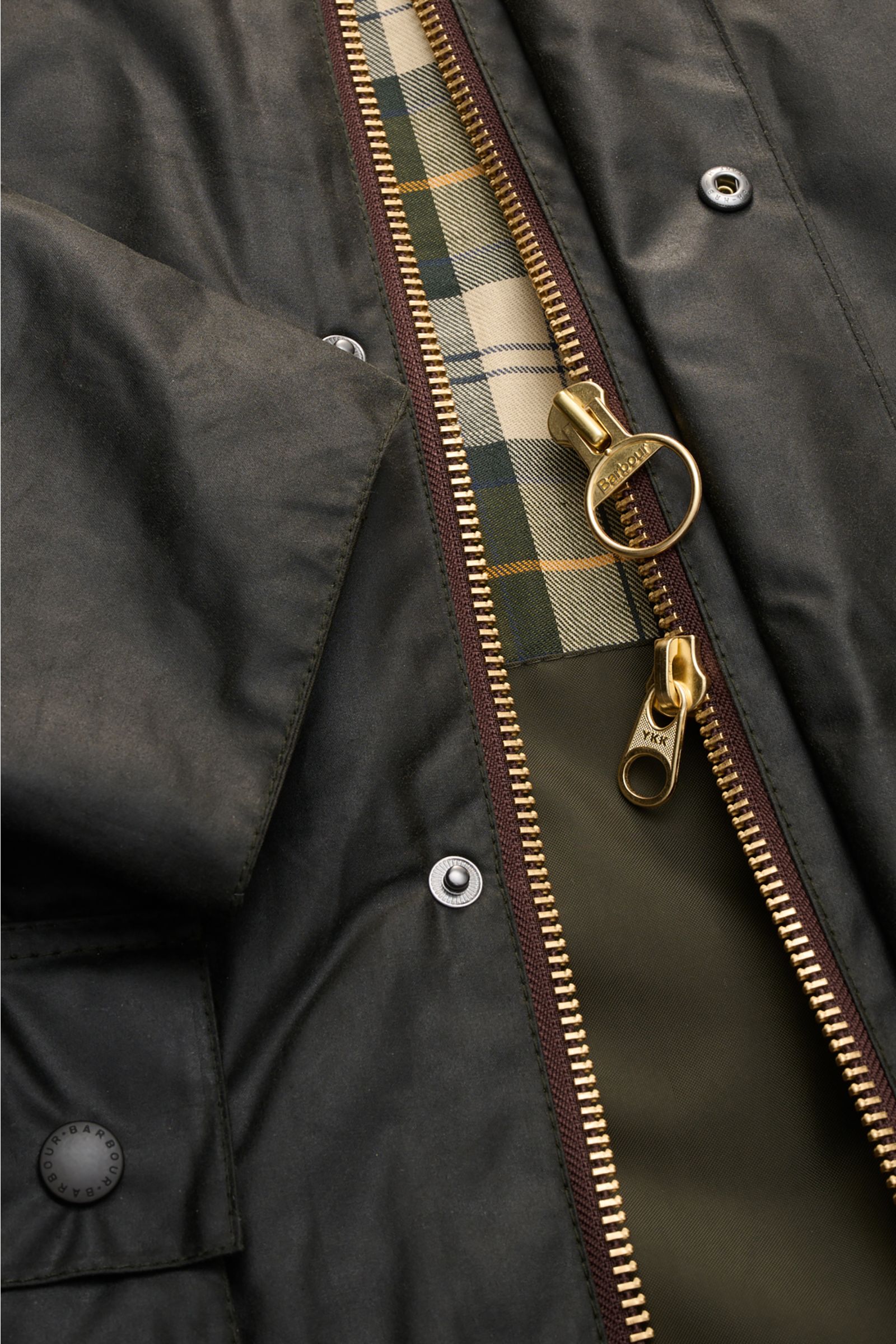 Close-up of the Barbour Waxed jacket 'Bedale' dark green showing brass two-way zipper, tartan lining, snap buttons, and fabric texture from top-down angle.

Description: Perfectly equipped for the changing seasons: The 'Bedale' from BARBOUR is made fro
