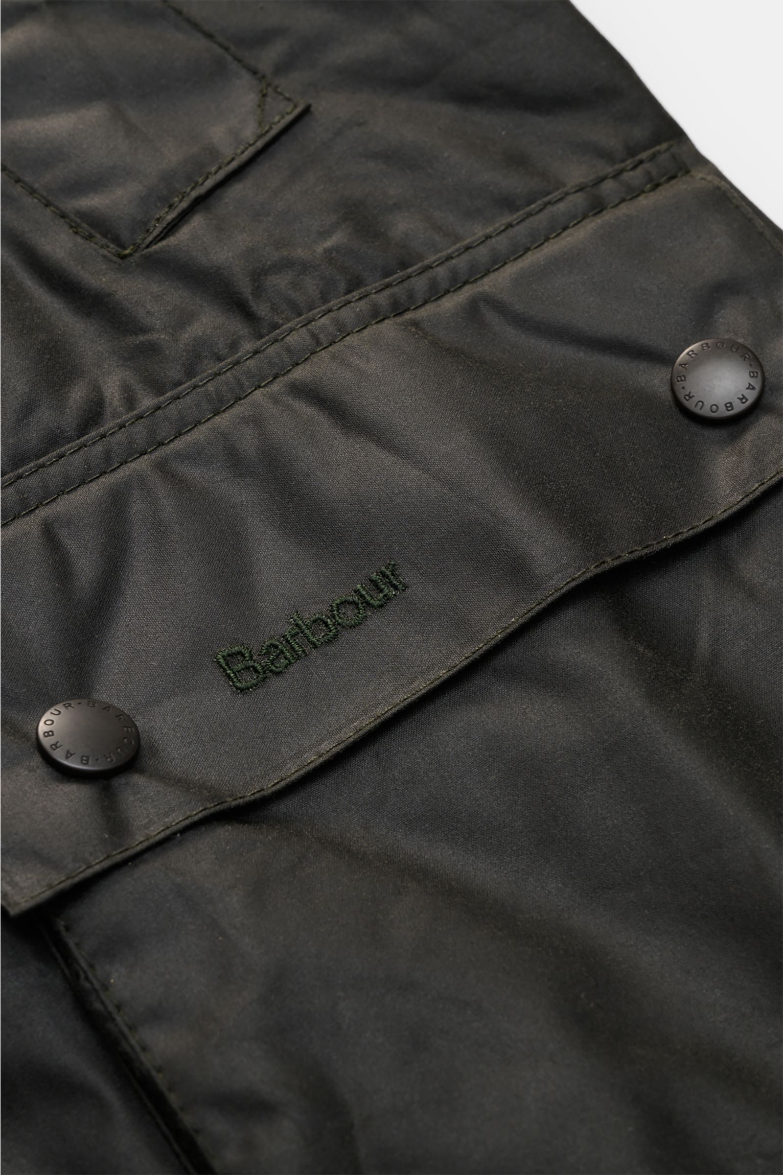 Barbour Wachsjacke 'Bedale' dunkelgrün, Nahaufnahme von gewachster Baumwolle mit Logostickerei auf der Patte und Druckknöpfen.