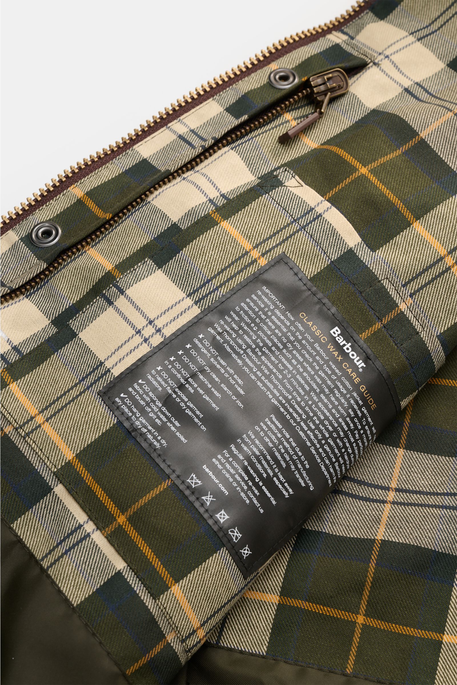 Innenseite der Barbour Wachsjacke 'Bedale' dunkelgrün mit typisch kariertem Tartan-Futter und Pflegehinweisen, Nahaufnahme, Blick von oben.
