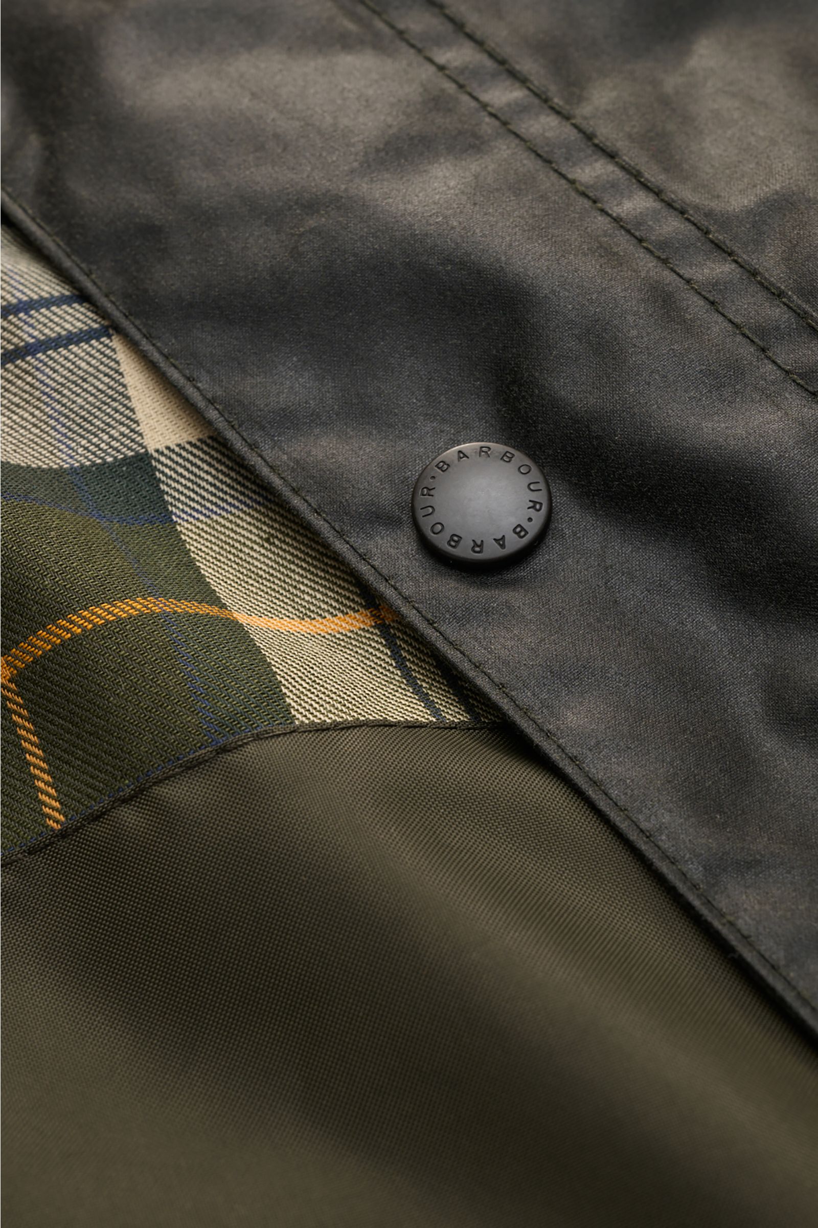 Close-up of Barbour Waxed jacket 'Bedale' dark green showing waxed cotton fabric, signature tartan lining, and black snap button with logo, photographed from a top-down angle. Perfectly equipped for the changing seasons: The 'Bedale' from BARBOUR is made 