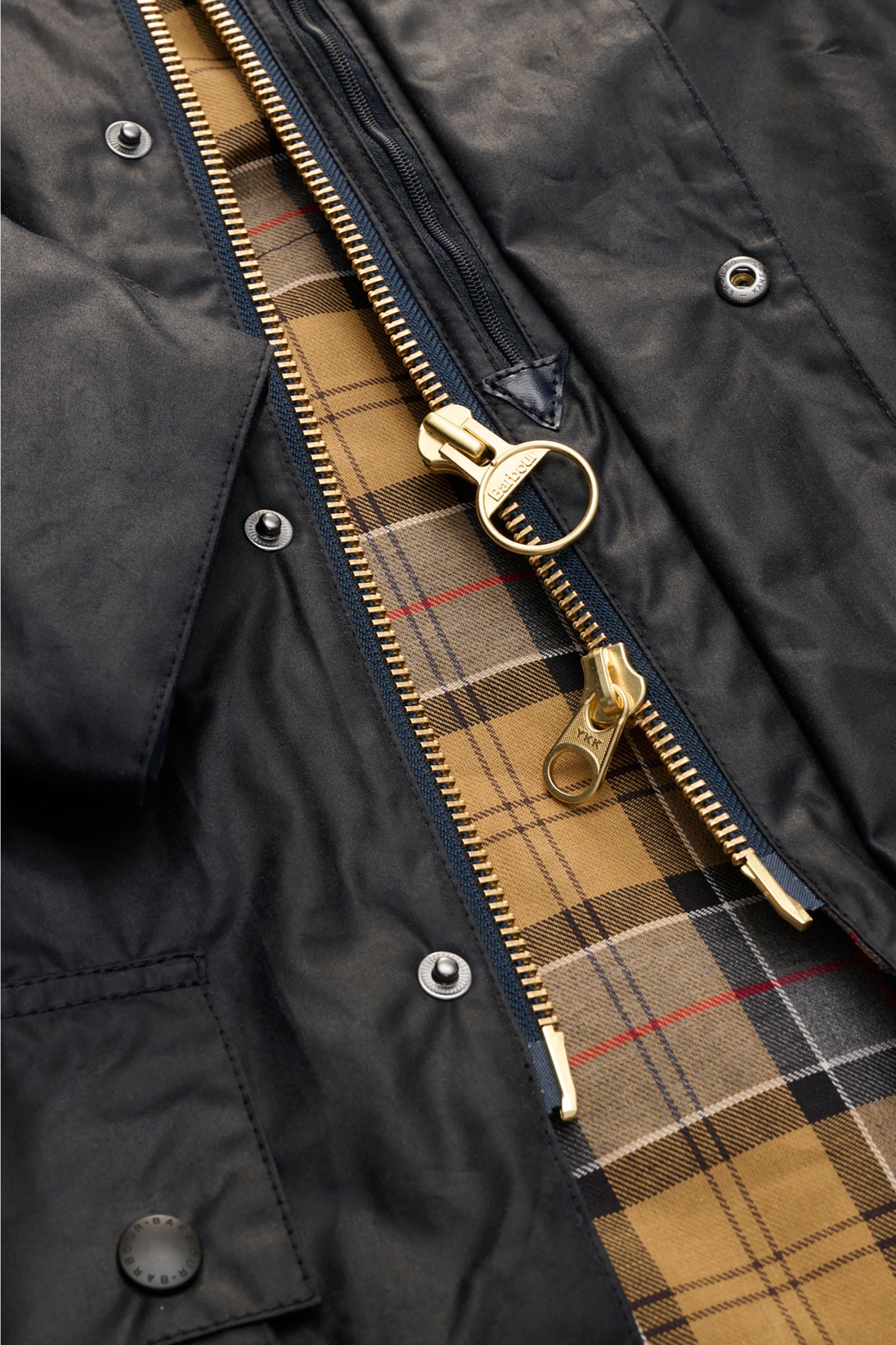 Close-up view of the Barbour Waxed jacket 'Beaufort' dark navy showing the robust waxed cotton fabric, gold-tone two-way zipper with ring pull, snap button storm flap, and classic tartan lining inside.

Description: A true classic in the British outdoor t