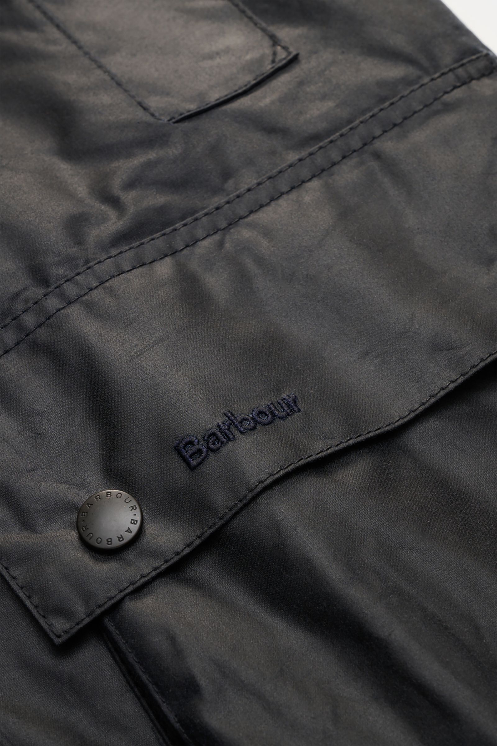 Nahaufnahme der Barbour Wachsjacke 'Beaufort' dark navy zeigt gewachste Baumwolle, Druckknopf und Logo auf der Fronttasche.