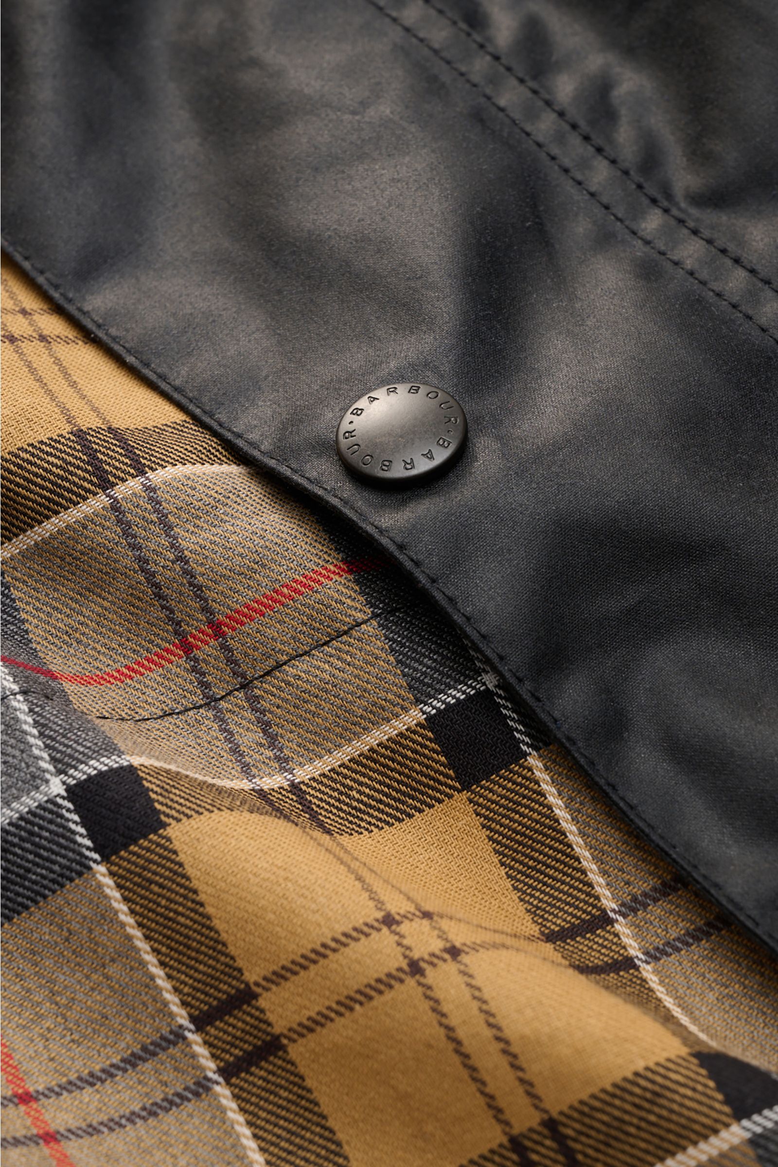 Close-up view of the Barbour Waxed jacket 'Beaufort' dark navy showing the black waxed cotton exterior with a branded snap button and classic tan tartan lining underneath.