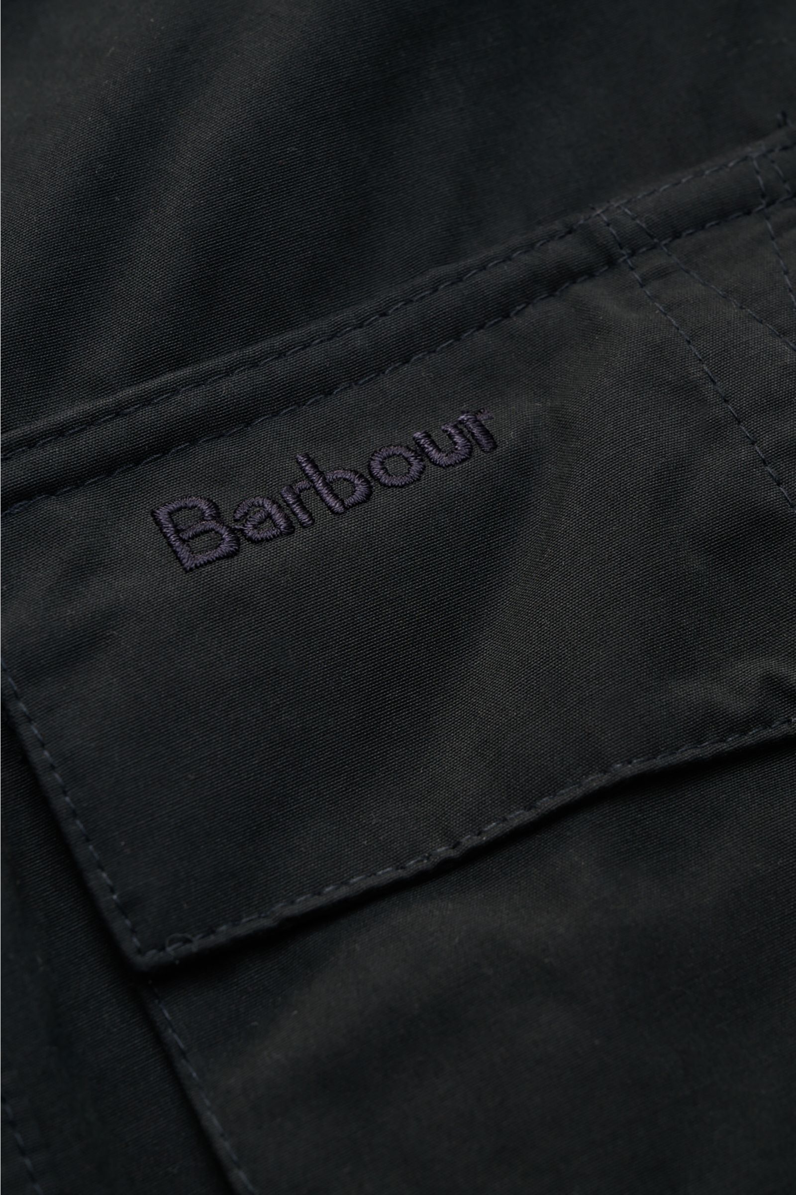 Close-up front pocket detail of Barbour Field jacket 'Sanderling' navy, showing the embroidered logo on cotton-microfibre fabric with a velvety feel, photographed from a top angle. Lightweight, functional, classic style, four bellows pockets, regular fit,
