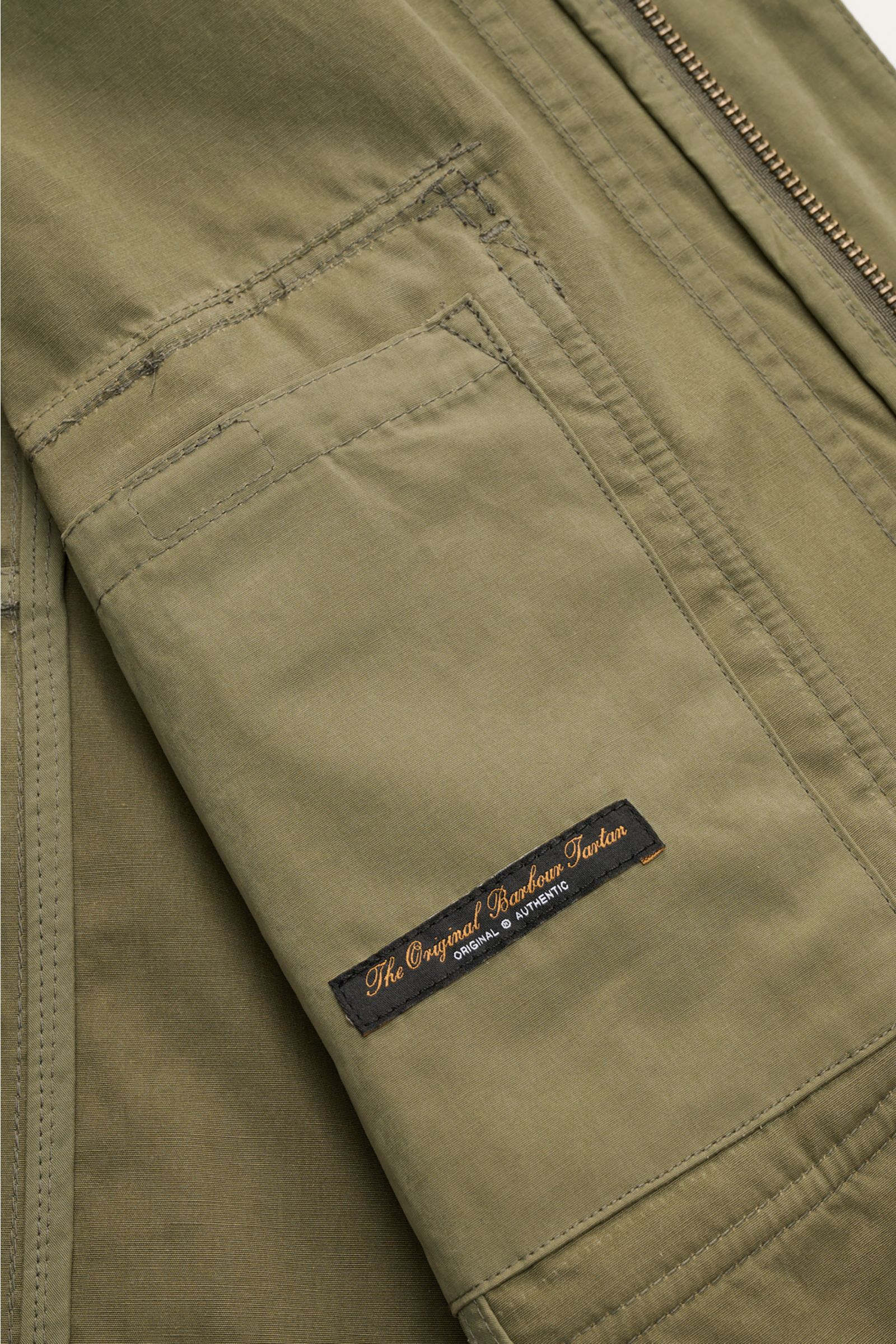 Close-up from above of the olive Barbour Field jacket 'Sanderling' showing lightweight fabric, sleeve pocket with Barbour Tartan label, and detailed stitching. Lightweight, functional and classic style: The versatile field jacket 'Sanderling' by BARBOUR i