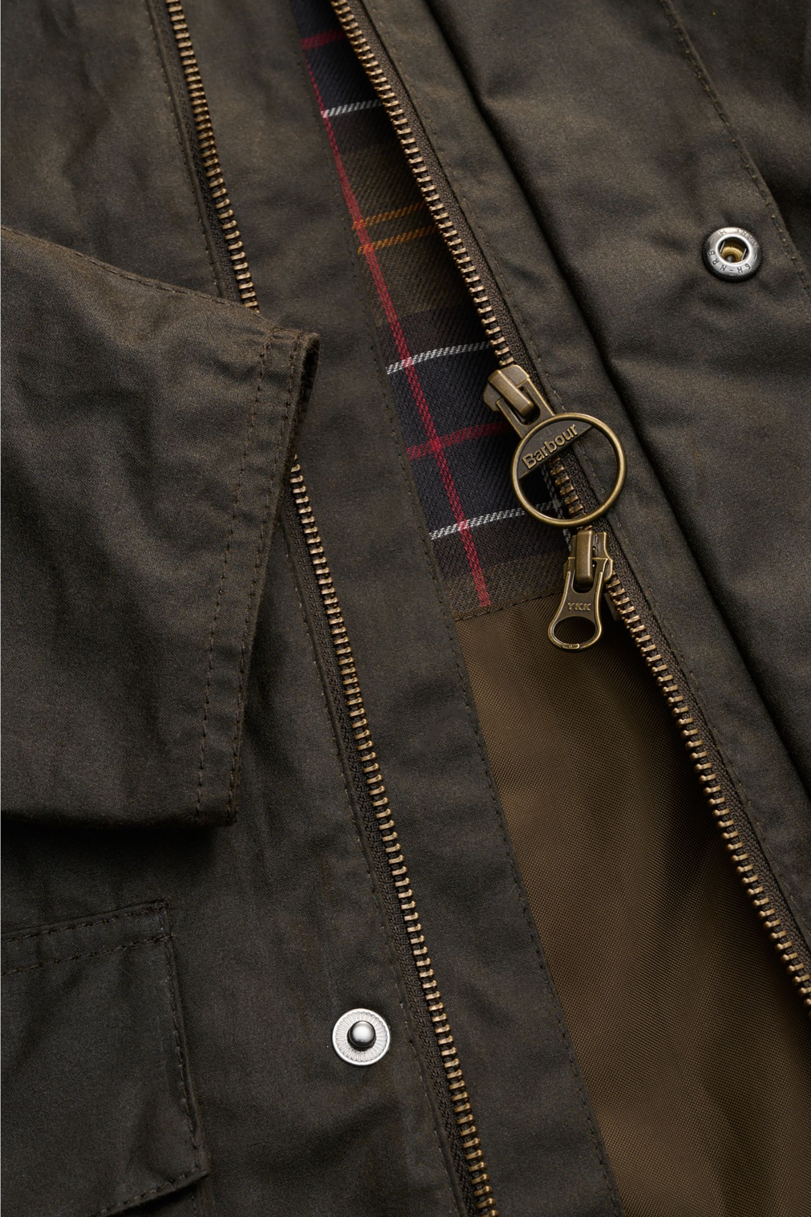 Close-up front view of the Barbour Waxed jacket 'Sander' dark olive showing the waxed cotton fabric, two-way brass zipper with ring pull, storm flap with snap buttons, and checked lining with corduroy-trimmed collar. Protection against wind and weather in