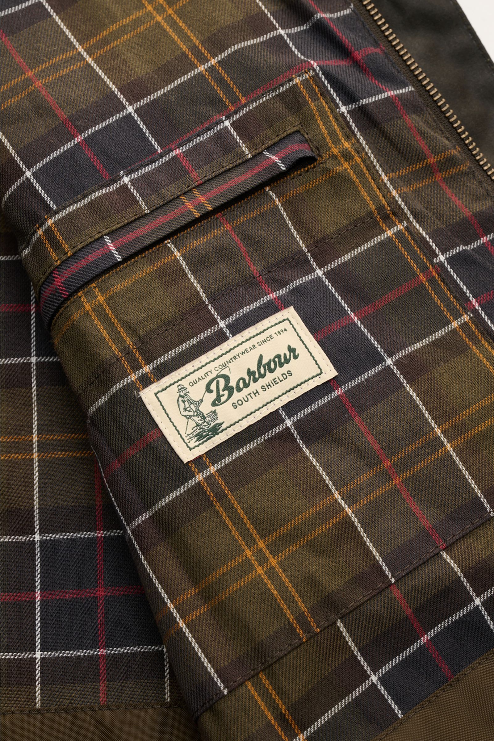 Close-up inside view of the checked lining and inside pocket with Barbour label of the Barbour Waxed jacket 'Sander' dark olive. 

Description: Protection against wind and weather in classic BARBOUR style: The waxed surface of the pure cotton jacket 'S