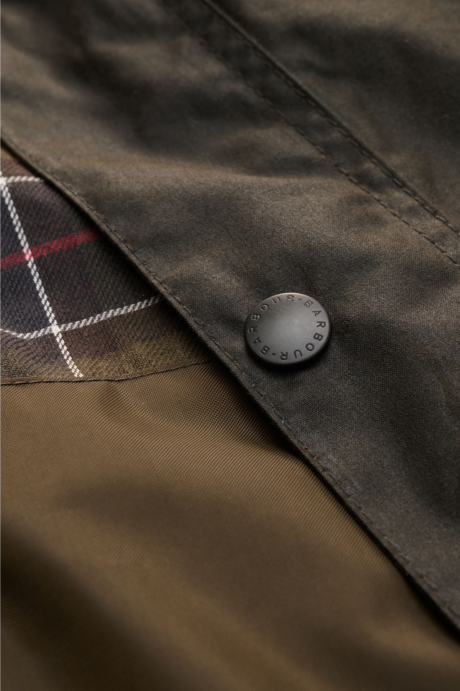 Close-up detail of the Barbour Waxed jacket 'Sander' dark olive, showing waxed cotton surface, corduroy collar trim, snap button, and checked lining from a top-down perspective.

Description: Protection against wind and weather in classic BARBOUR style
