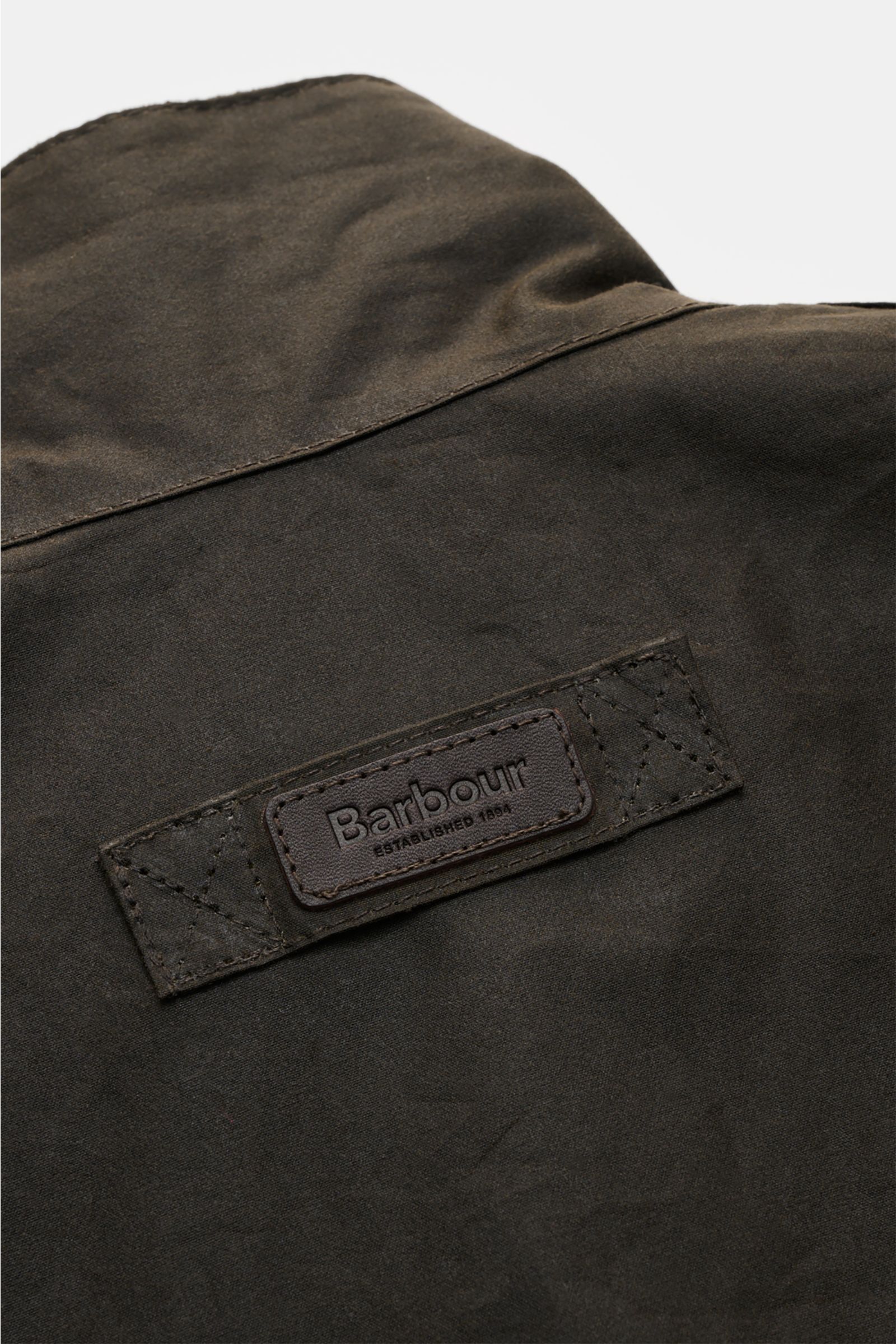 Close-up image of the back collar area of the Barbour Waxed jacket 'Sander' dark olive, showcasing the leather Barbour logo patch. Protection against wind and weather in classic BARBOUR style: The waxed surface of the pure cotton jacket 'Sander' guarantee