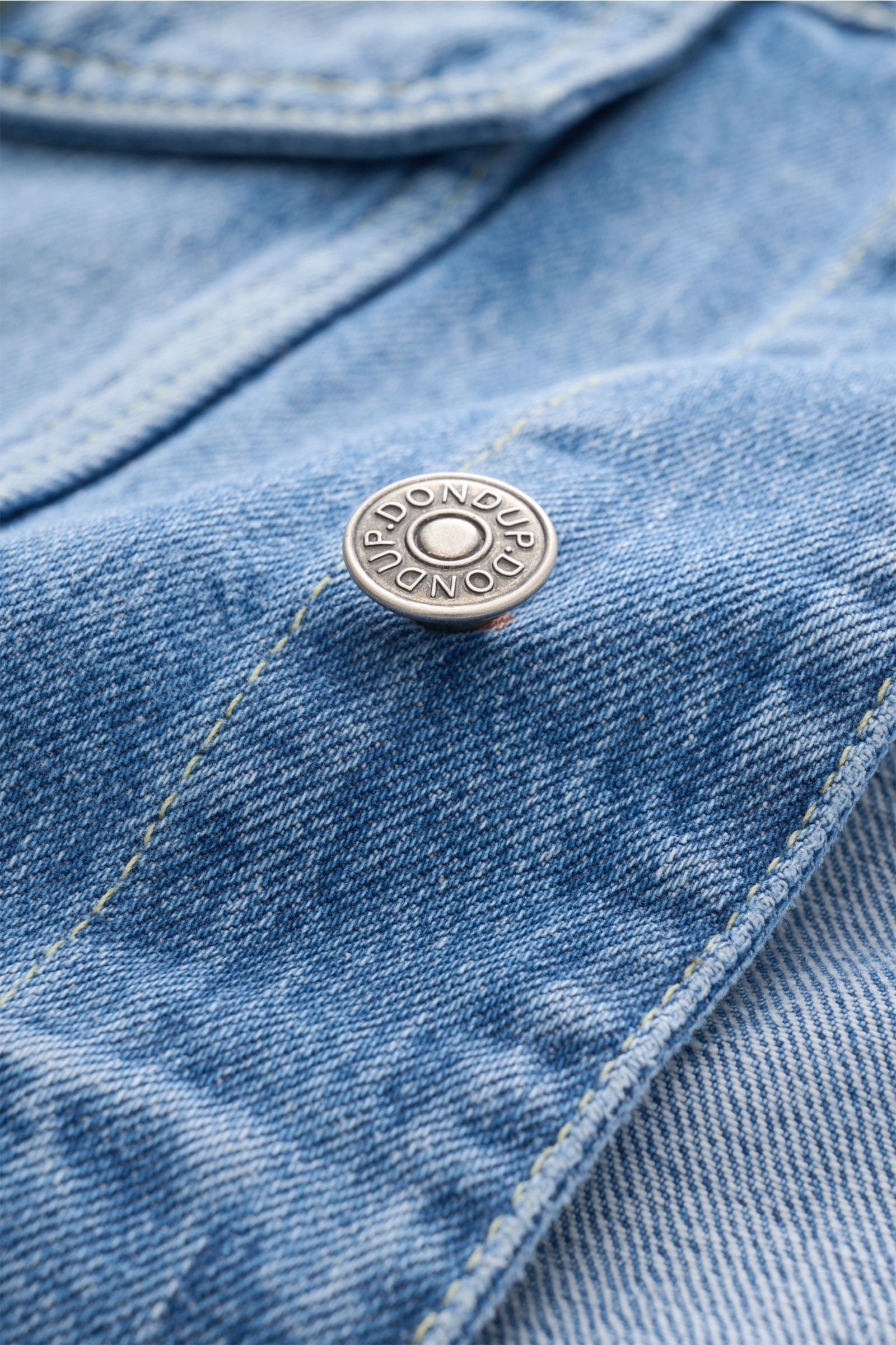 Nahaufnahme einer Metall-Knopfleiste mit Dondup-Logo auf der rauchblauen Dondup Jeansjacke, Slim Fit aus reiner Baumwolle, mit Kontrastnähten.