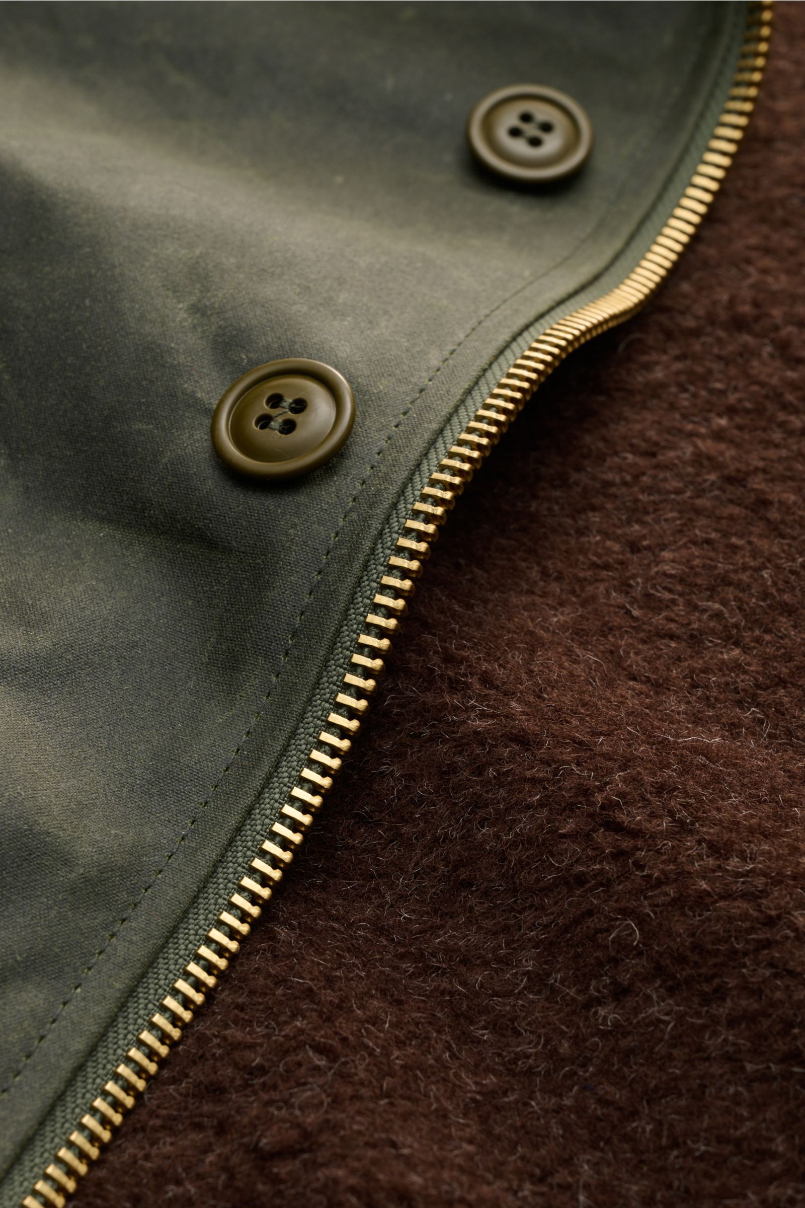Manifattura Ceccarelli Wachsjacke 'Deck Jacket' dunkelgrün in Nahaufnahme, zeigt gewachste Baumwolle, goldenen Reißverschluss, Knöpfe und braunes Alpakawollfutter.