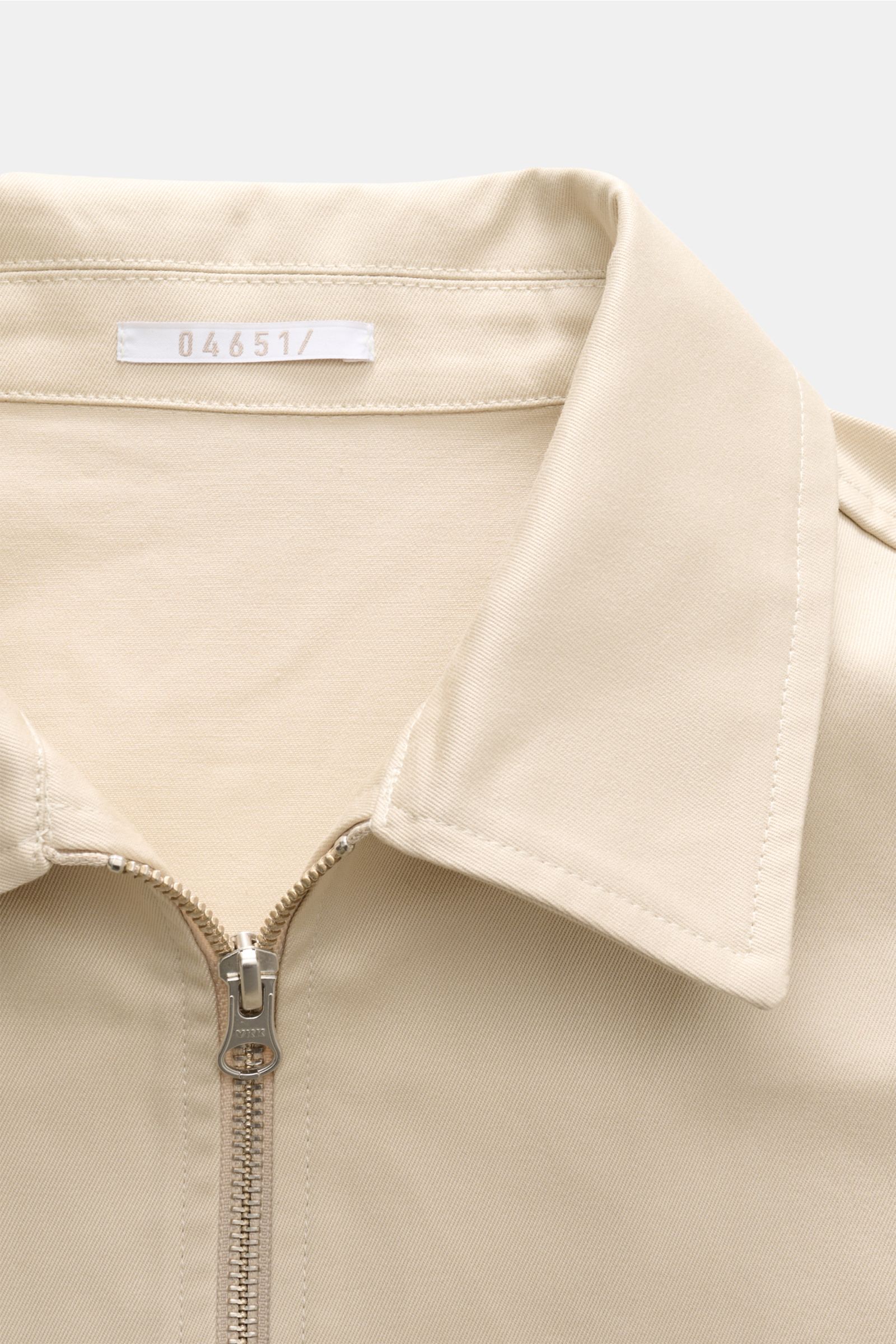 Close-up image of the collar and zipper front of the 04651/ A trip in a bag Blouson 'Griffin' beige, photographed from above, showcasing its pure cotton fabric, soft touch, slim fit, robust zipper, turn-down collar, piped front pockets, elastic hem and sl