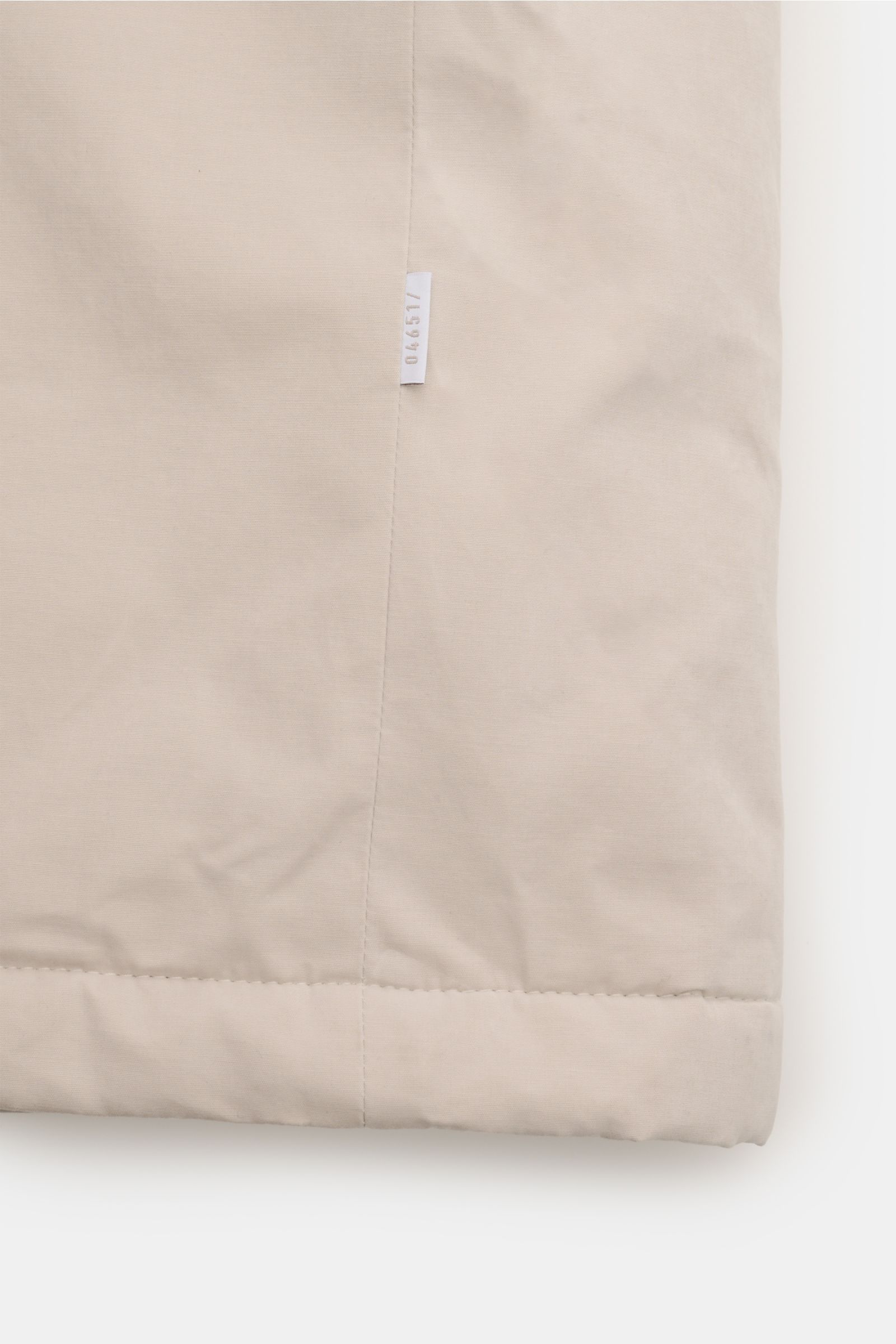 Close-up view of the hem and side seam of the 04651/ A trip in a bag Parka 'MC26' sand, made from lightly waxed cotton with a soft touch, featuring a small white fabric label with '04651' printed on it. Inspiriert von den Olympischen Winterspielen in Cort