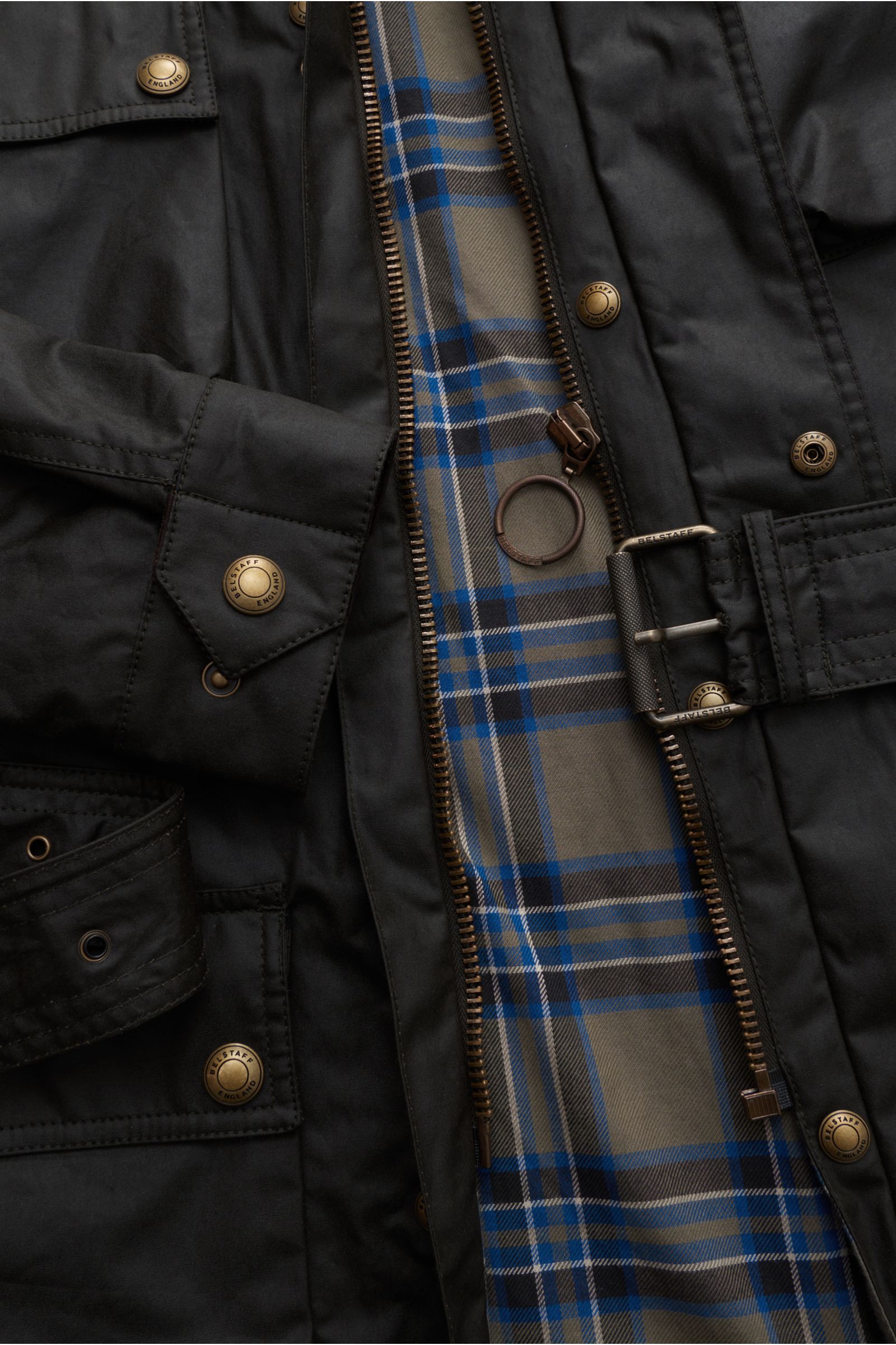 Close-up top-down view of the black Belstaff Wachsjacke 'Trialmaster' showing waxed cotton fabric, bronze buttons, zipper, belt buckle, and blue plaid lining.
