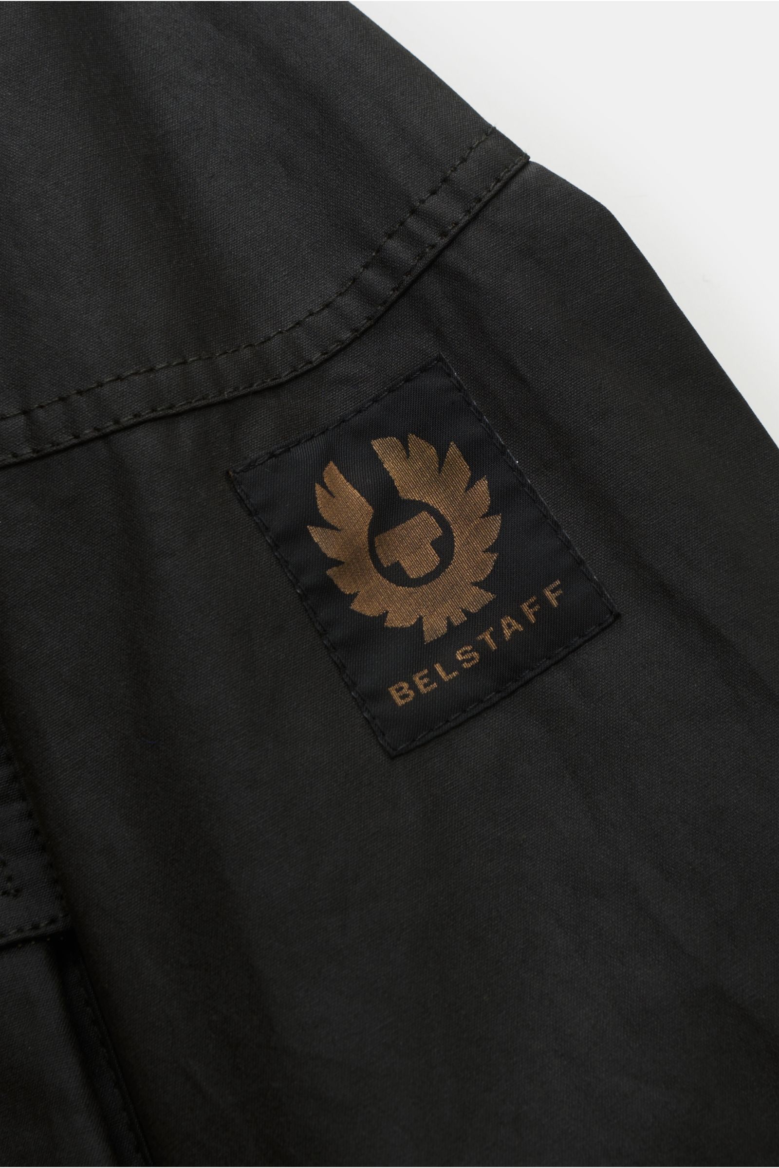 Close-up of the Belstaff Wachsjacke 'Trialmaster' schwarz showing the BELSTAFF logo patch on the upper arm, photographed from a side angle. Strapazierfähig, funktional und zeitlos – die legendäre Jacke 'Trialmaster' mit charakteristischem Taillengürtel un