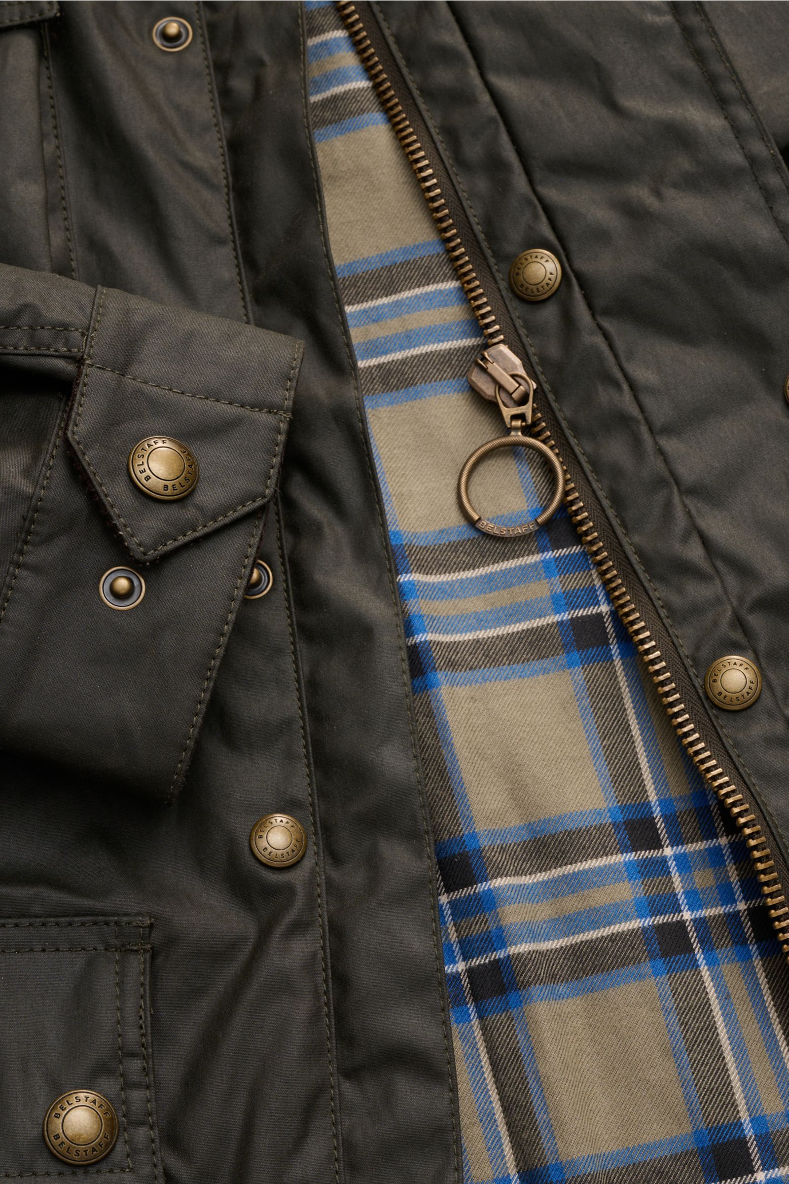 Nahaufnahme der Belstaff Wachsjacke 'Trialmaster' dark olive von vorne, gewachste Baumwolle, robuster Reißverschluss mit Ring, aufgesetzte Taschen, innen kariertes Futter.