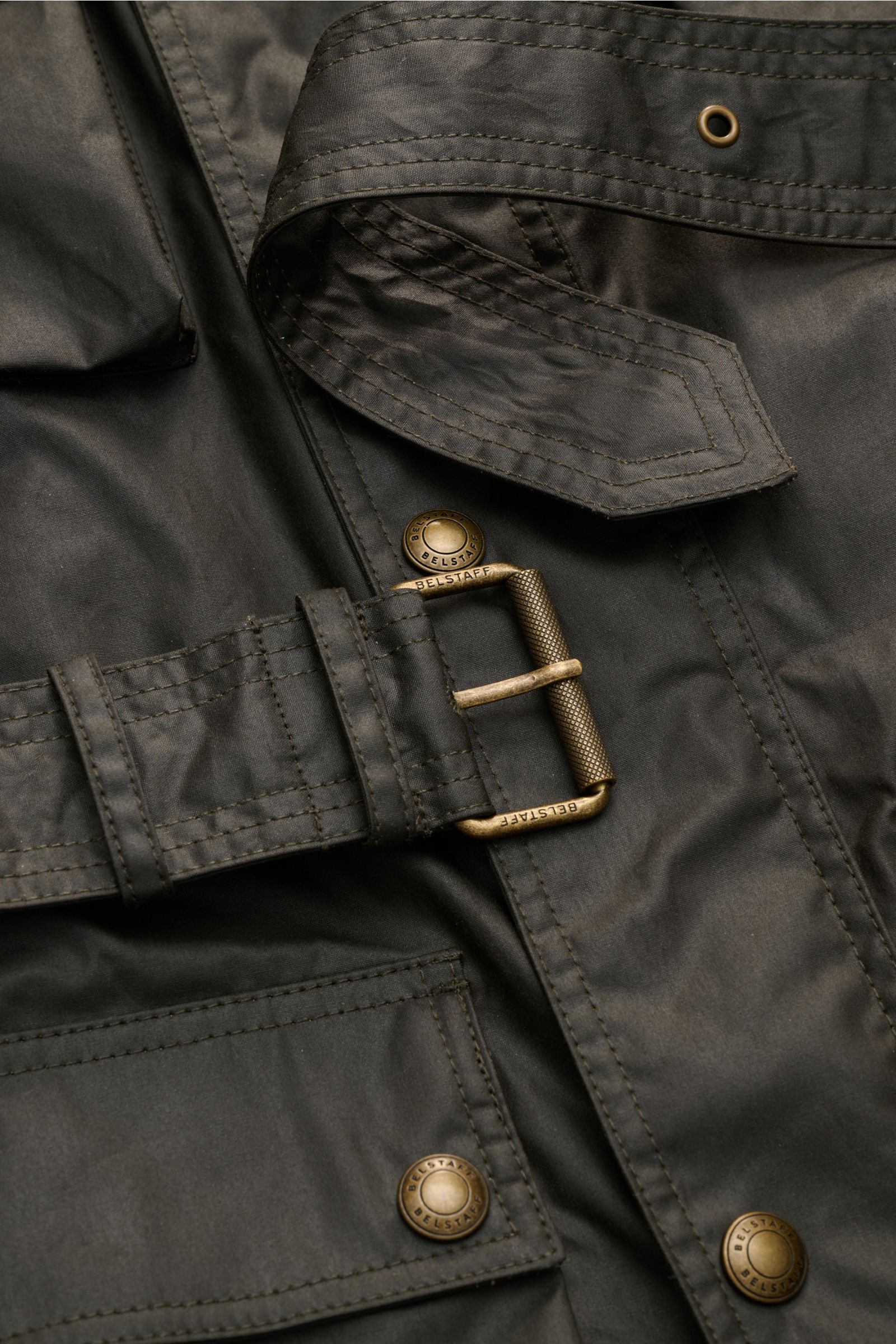 Nahaufnahme der Belstaff Wachsjacke 'Trialmaster' dark olive, Detailansicht von Taillengürtel mit Dornschließe und aufgesetzter Tasche aus gewachster, wasserabweisender Baumwolle.