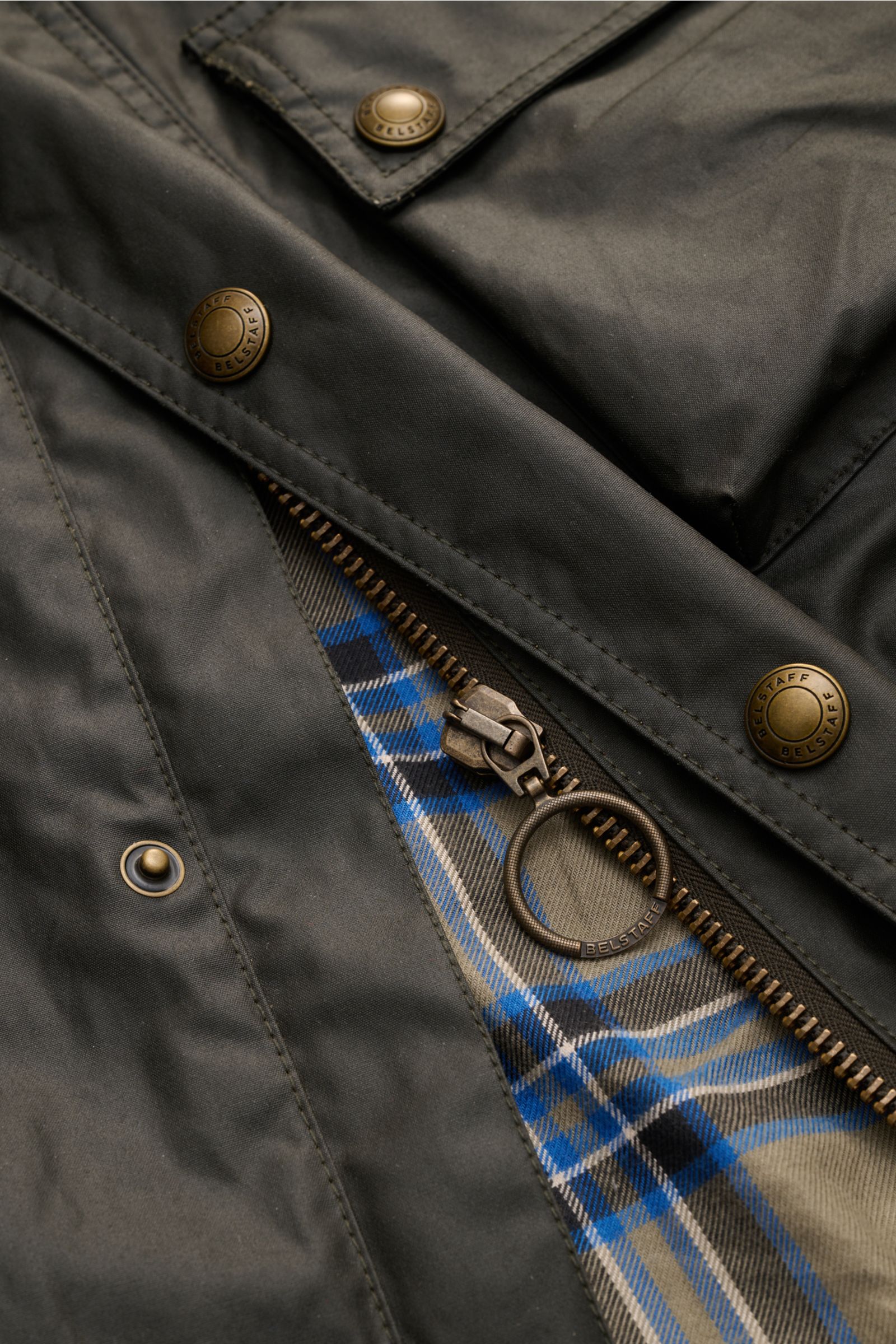 Nahaufnahme der Belstaff Wachsjacke 'Racemaster' dark olive aus gewachster Baumwolle, Fokus auf Reißverschluss mit Ring und karierter Innenfutterperspektive.