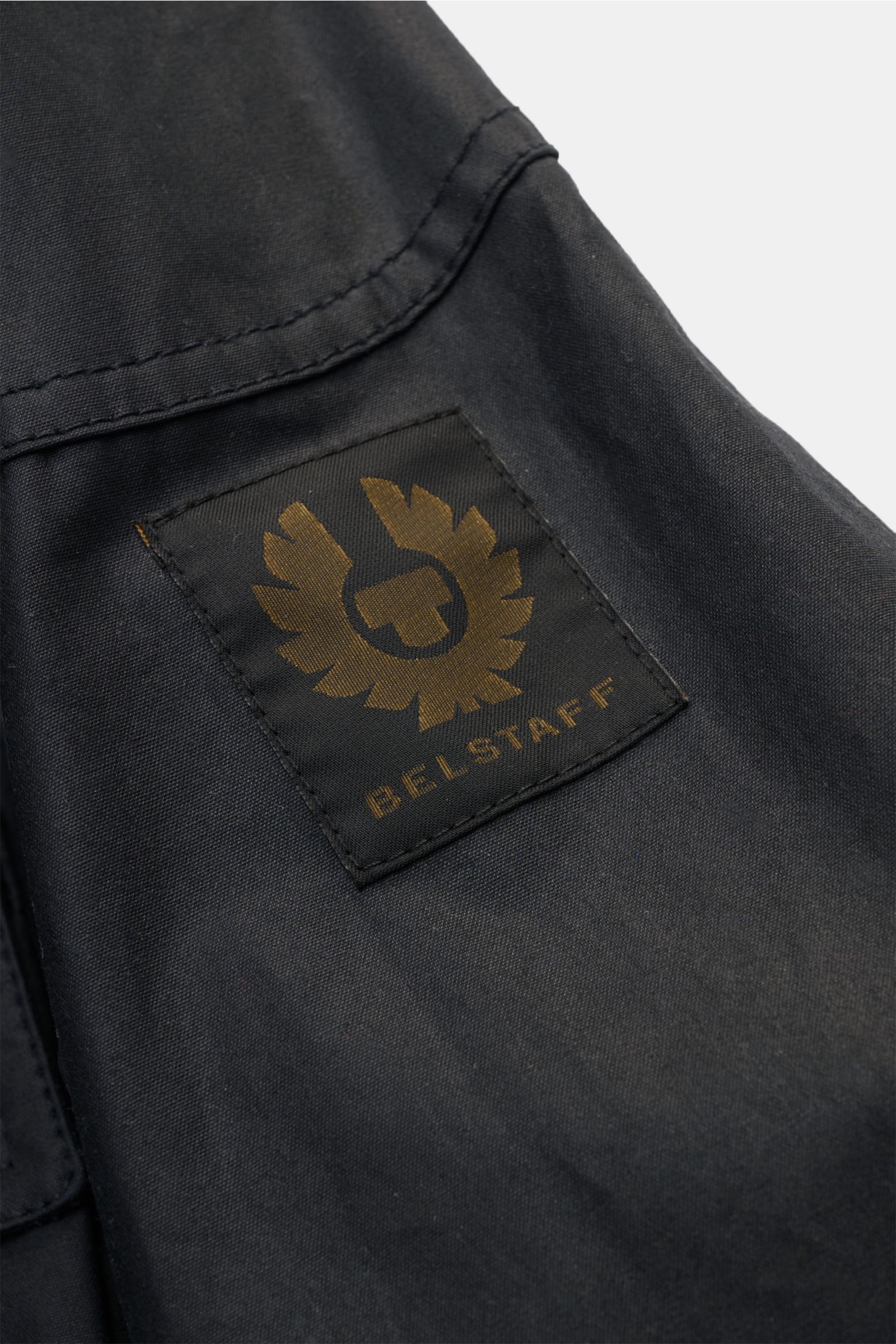Nahaufnahme der Belstaff Wachsjacke 'Fieldmaster' dark navy, Fokus auf gewachste Baumwolle und das BELSTAFF-Weblabel am Ärmel.