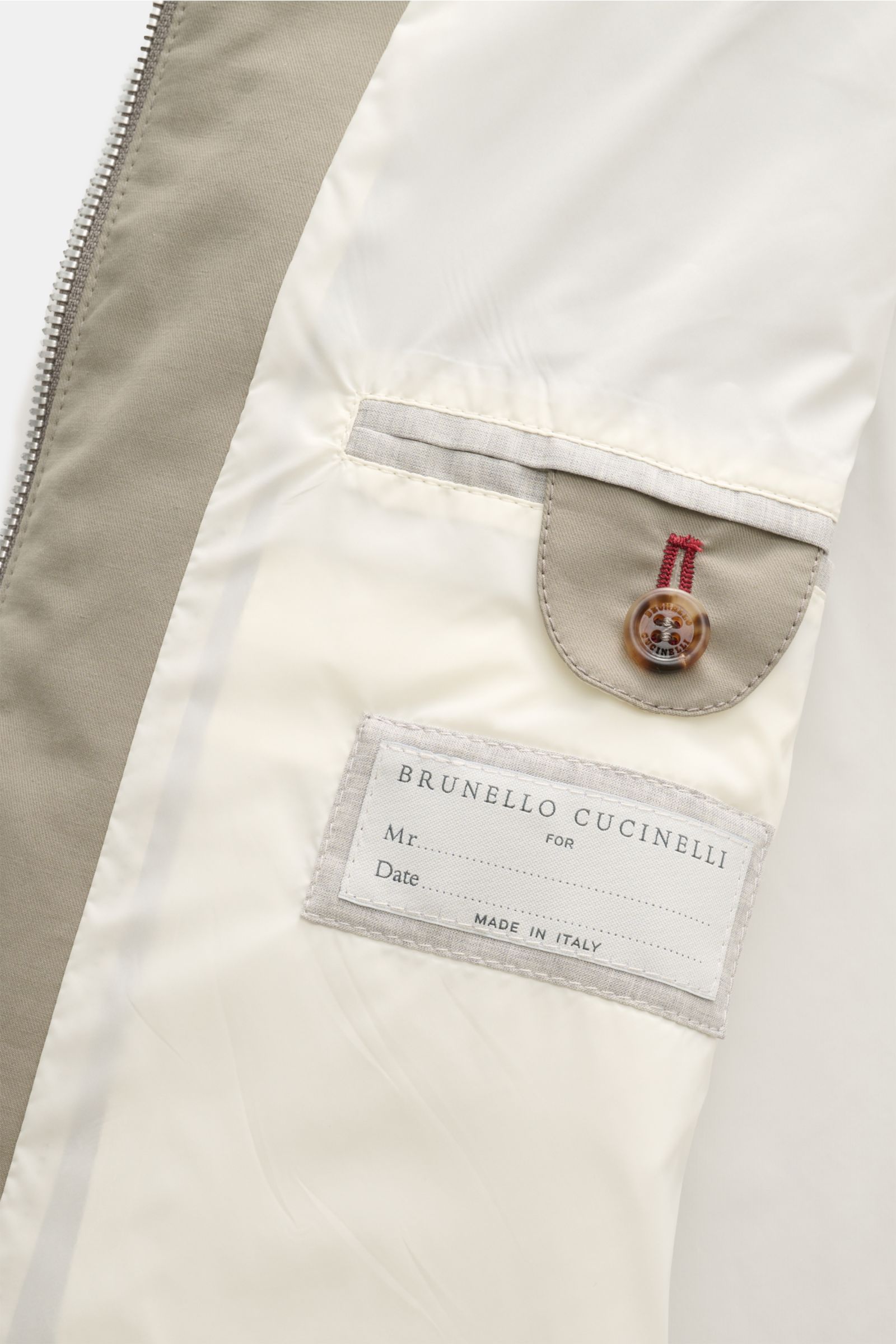 Close-up inside view of the Brunello Cucinelli Blouson salbei showing the cream lining, buttoned pocket flap with red stitching, and label tag.