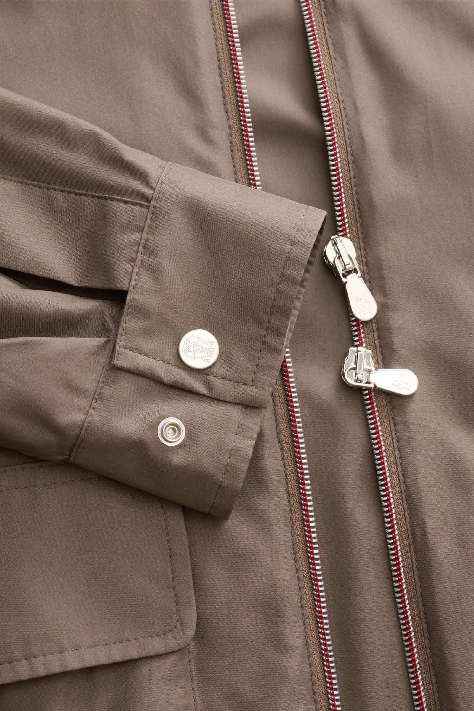 Close-up front view of the taupe Brunello Cucinelli Jacke showing slim fit sleeve cuff with branded buttons and two-way zipper with red trim, luxurious silk-microfiber blend, smooth touch, unconstructed design, flap pockets with snap buttons, and turn-dow