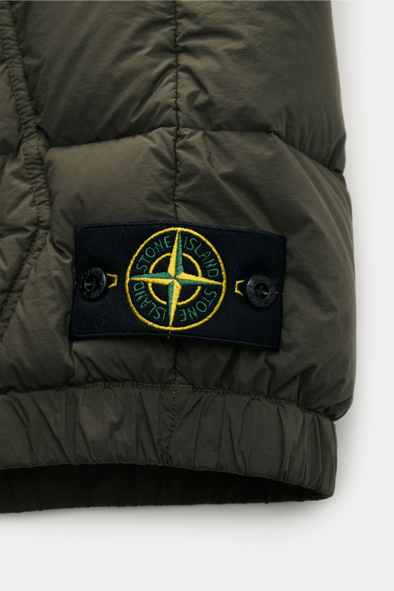 Close-up of the hem and signature compass badge on the Stone Island Down gilet 'Seamless Tunnel Nylon Down-TC' olive, photographed from the side to highlight the elasticated hem and fabric texture. 

The outdoor gilet 'Seamless Tunnel Nylon Down-TC' by