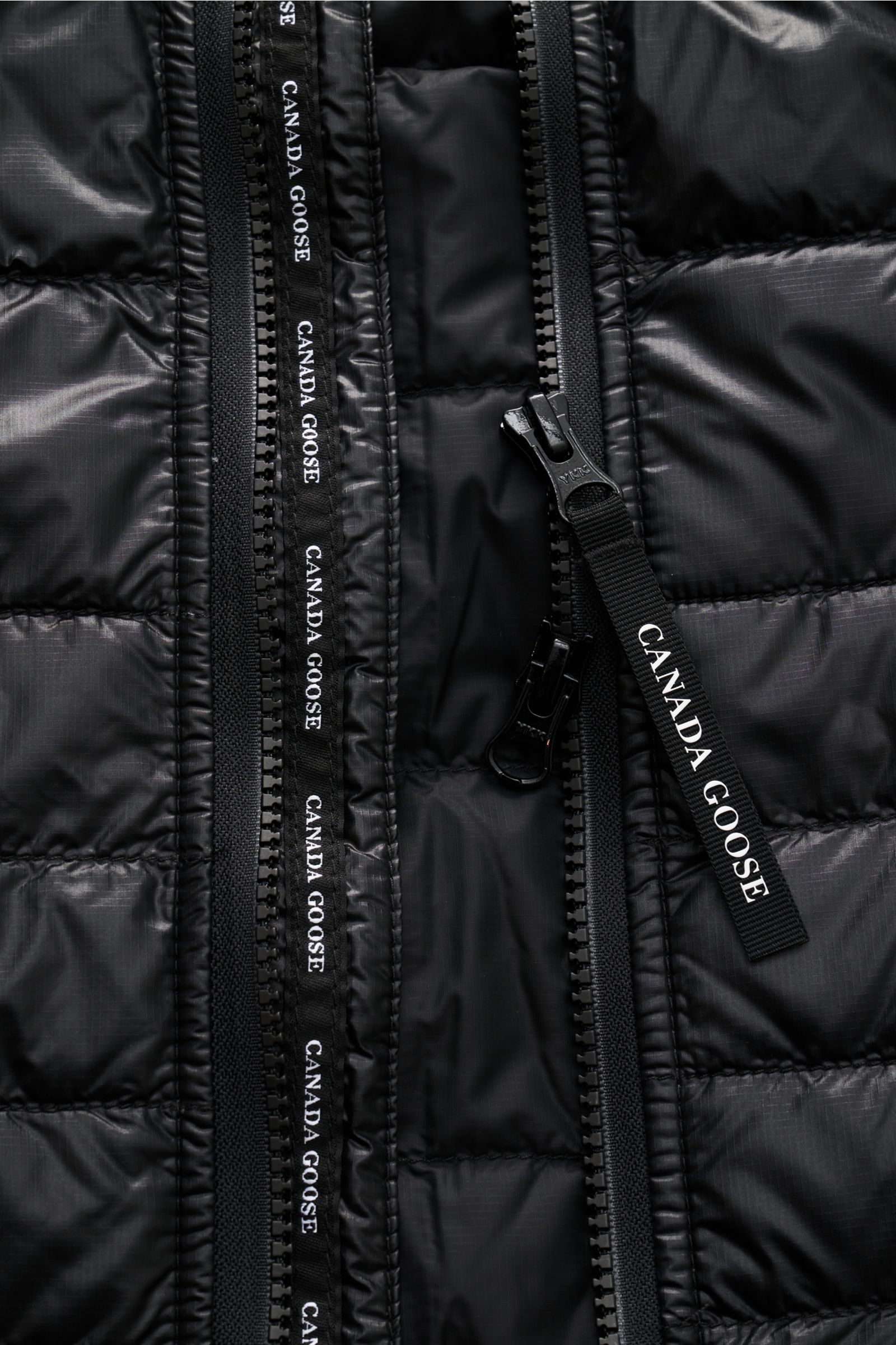 Nahaufnahme der schwarzen Canada Goose Daunenweste 'Hybridge Lite' mit Ripstop-Microfaser, Reißverschluss und Logo-Zipper von vorne.