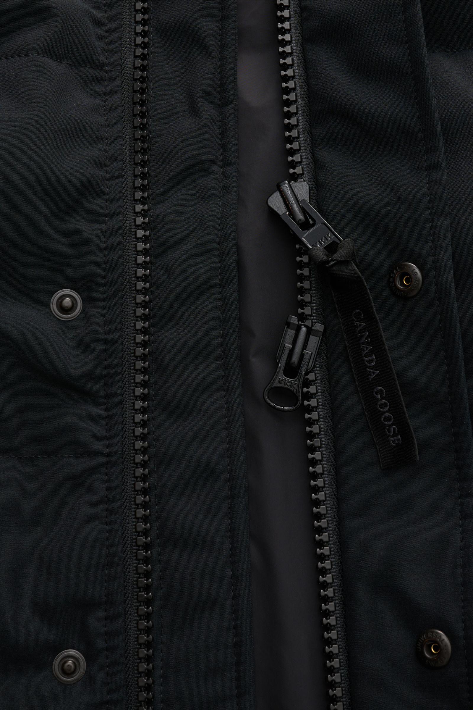 Nahaufnahme der Canada Goose Daunenweste 'Garson' schwarz, Fokus auf robustem Zwei-Wege-Reißverschluss mit CANADA GOOSE-Schleife, Baumwoll-Microfaser-Mix, wasserabweisend, Slim Fit, Stehkragen und Druckknöpfen sichtbar.