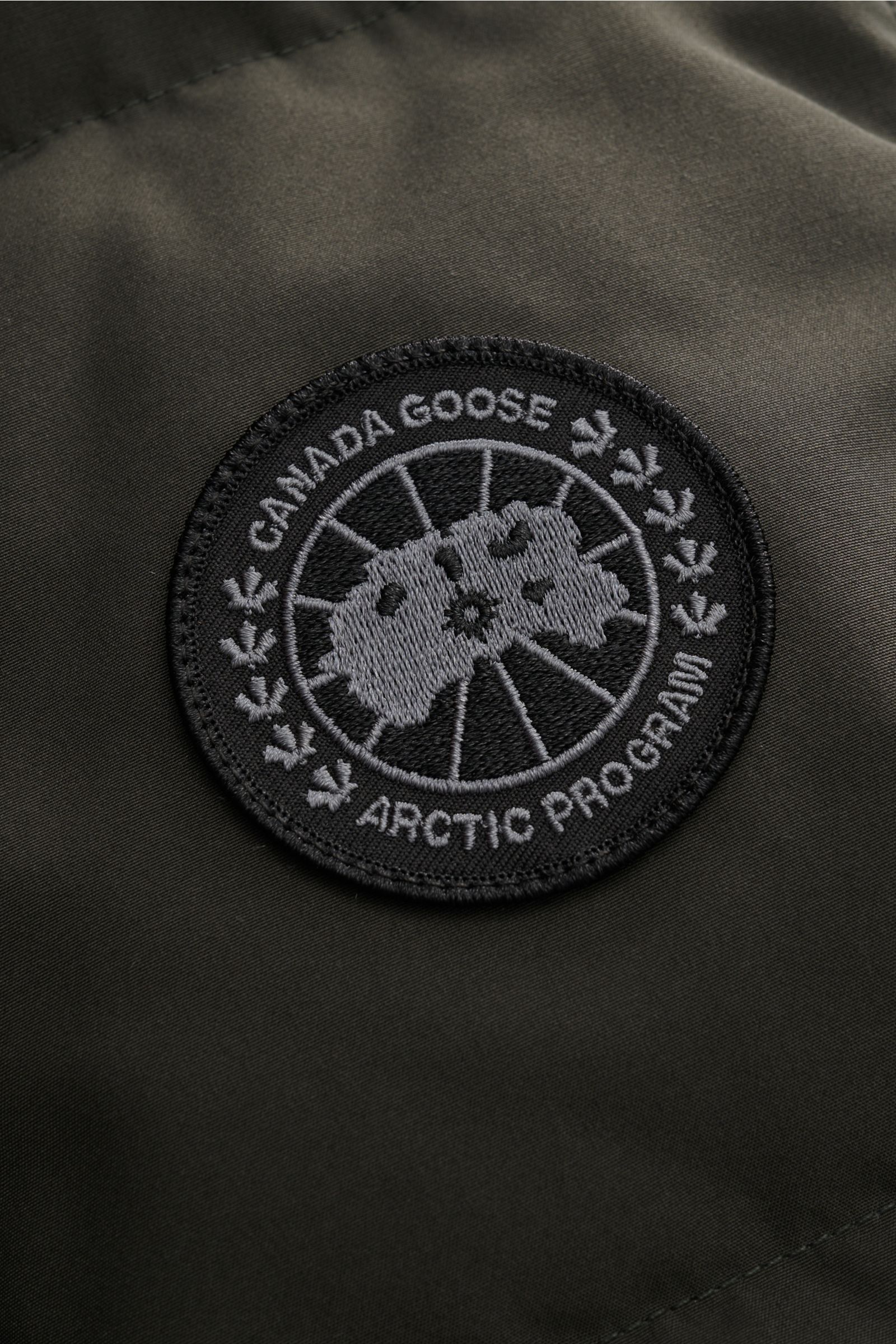 Canada Goose Daunenweste 'Garson' graugrün im Detail, Nahaufnahme des wasserabweisenden Materials mit CANADA GOOSE-Weblabel, Mix aus Baumwolle und Microfaser.