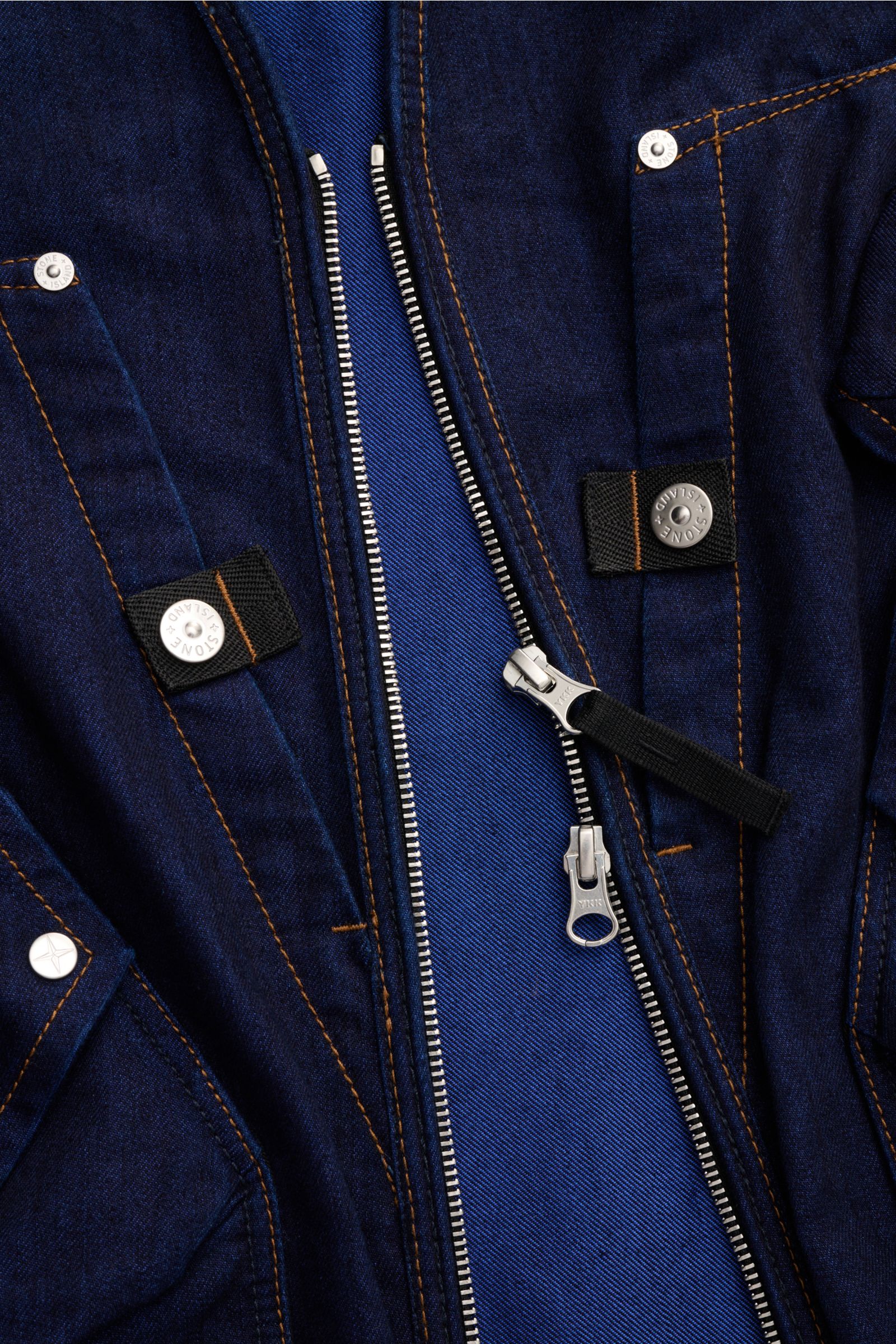 Nahaufnahme der Stone Island Jeansweste 'Indigo Polypropylene Denim Light' dunkelblau im Utility-Look mit robustem Zwei-Wege-Reißverschluss, fünf Fronttaschen und markentypischem Kompass-Badge, fotografiert aus schräger Perspektive.