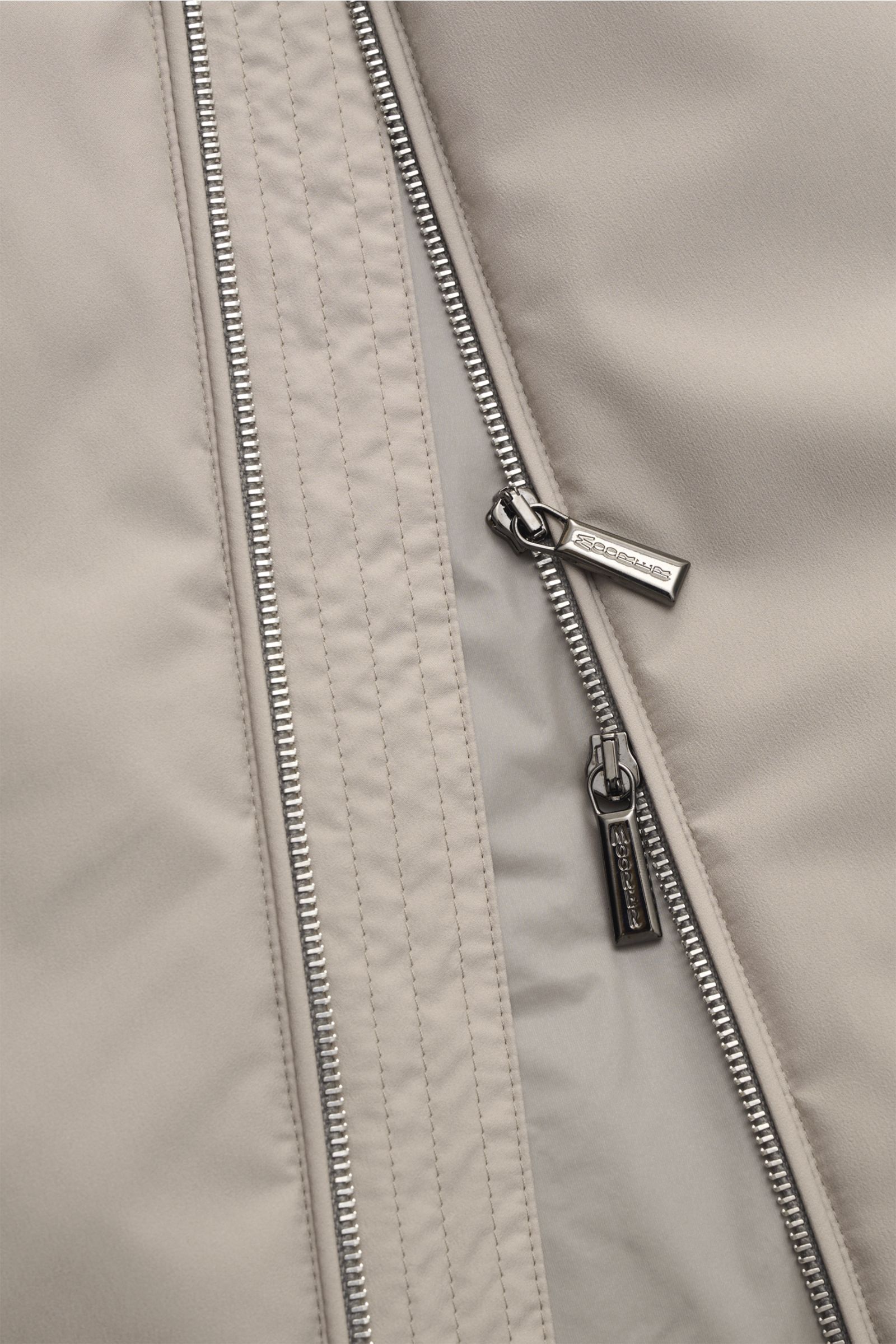 Close-up front view of the beige Moorer Outdoor-Weste 'Senio-Skt' showing its water-repellent microfiber fabric, smooth texture, slim fit, two-way zipper with branded metal pulls, narrow stand-up collar, and angled zipper front pockets. Die leichte Out