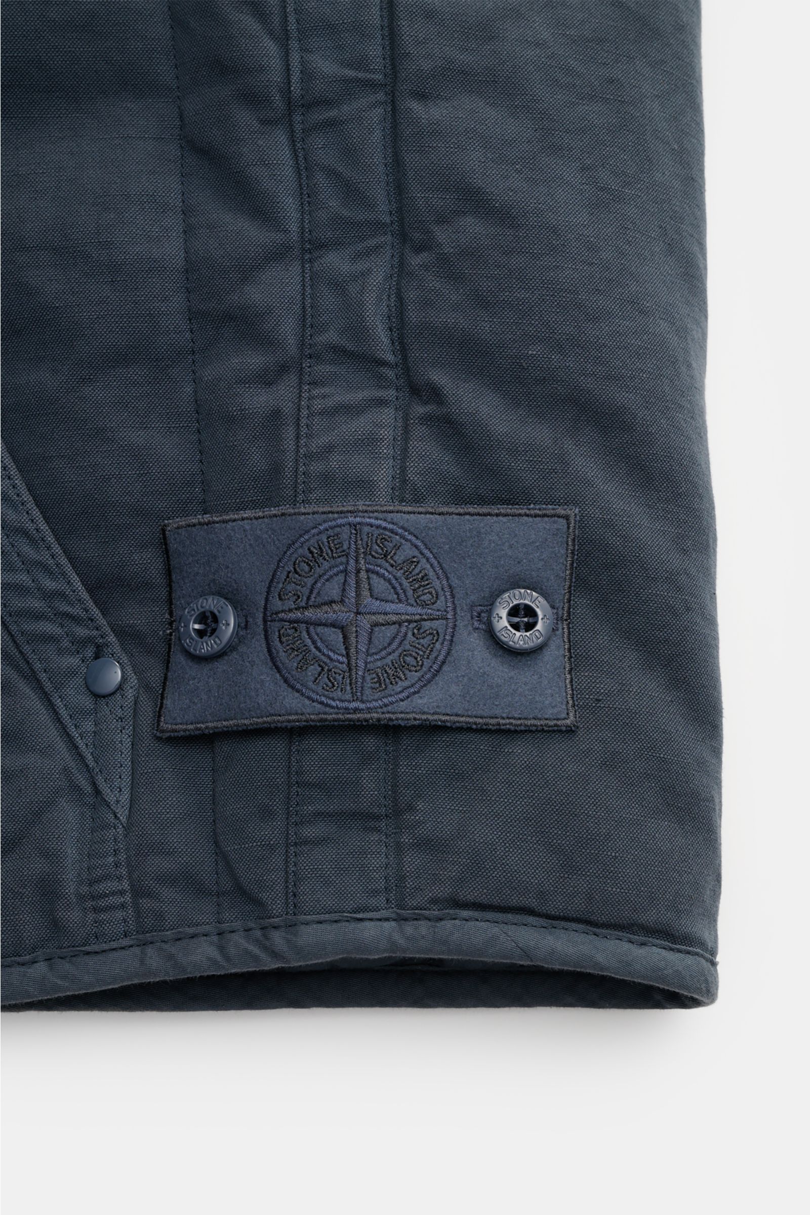 Nahaufnahme der Stone Island Outdoorweste 'Ghost Piece' graublau aus Baumwolle-Leinen mit tonalem Badge am Saum, Rippbündchen und Slim Fit.
