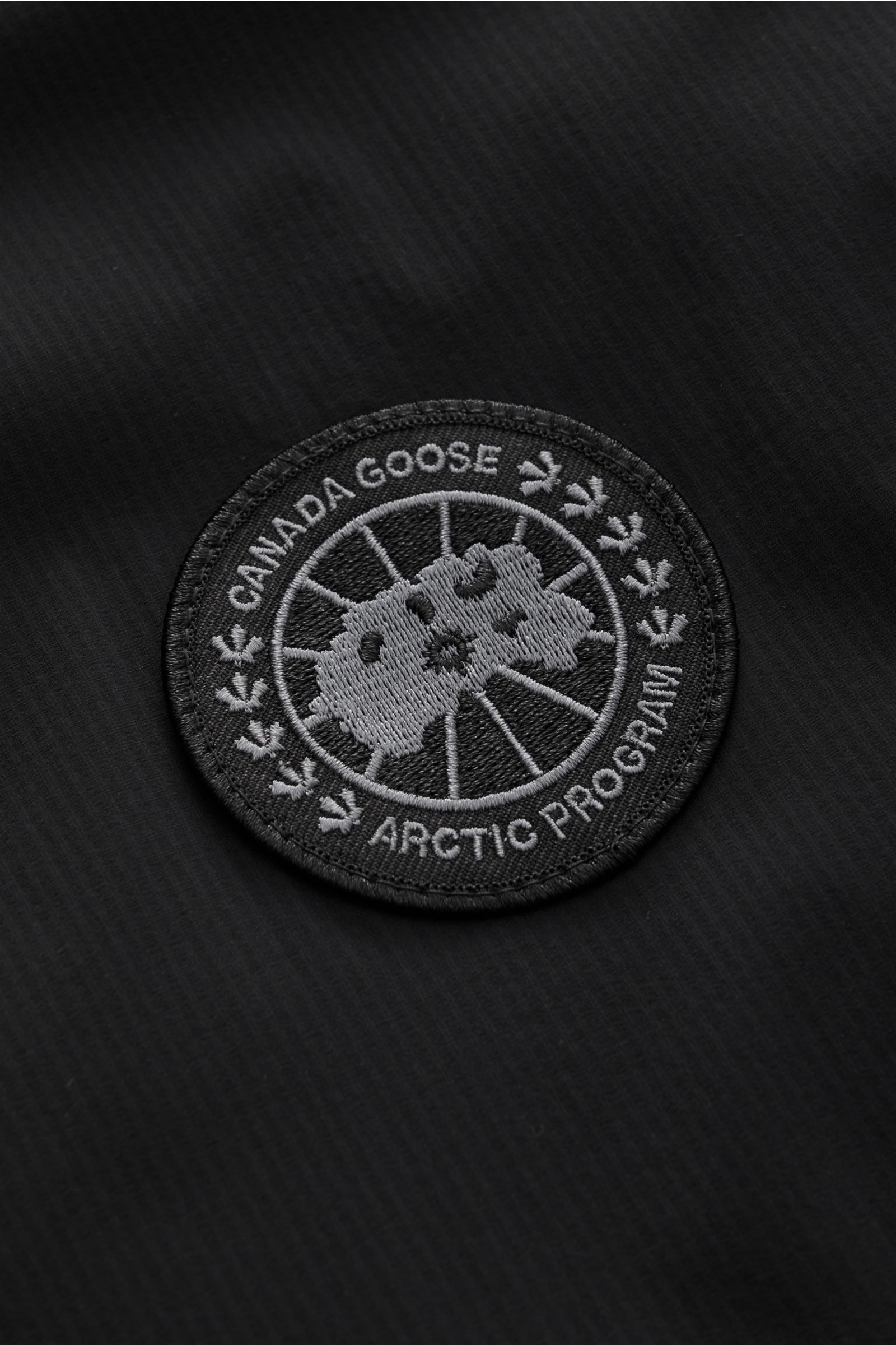 Canada Goose Daunenweste 'Lodge' schwarz aus superleichtem Ripstop-Microfaser mit wind- und wasserabweisendem Stoff, Slim Fit, Stehkragen, Zwei-Wege-Reißverschluss, tonalem Weblabel und regulierbarem Saum, Detailaufnahme des Logos auf der Front.