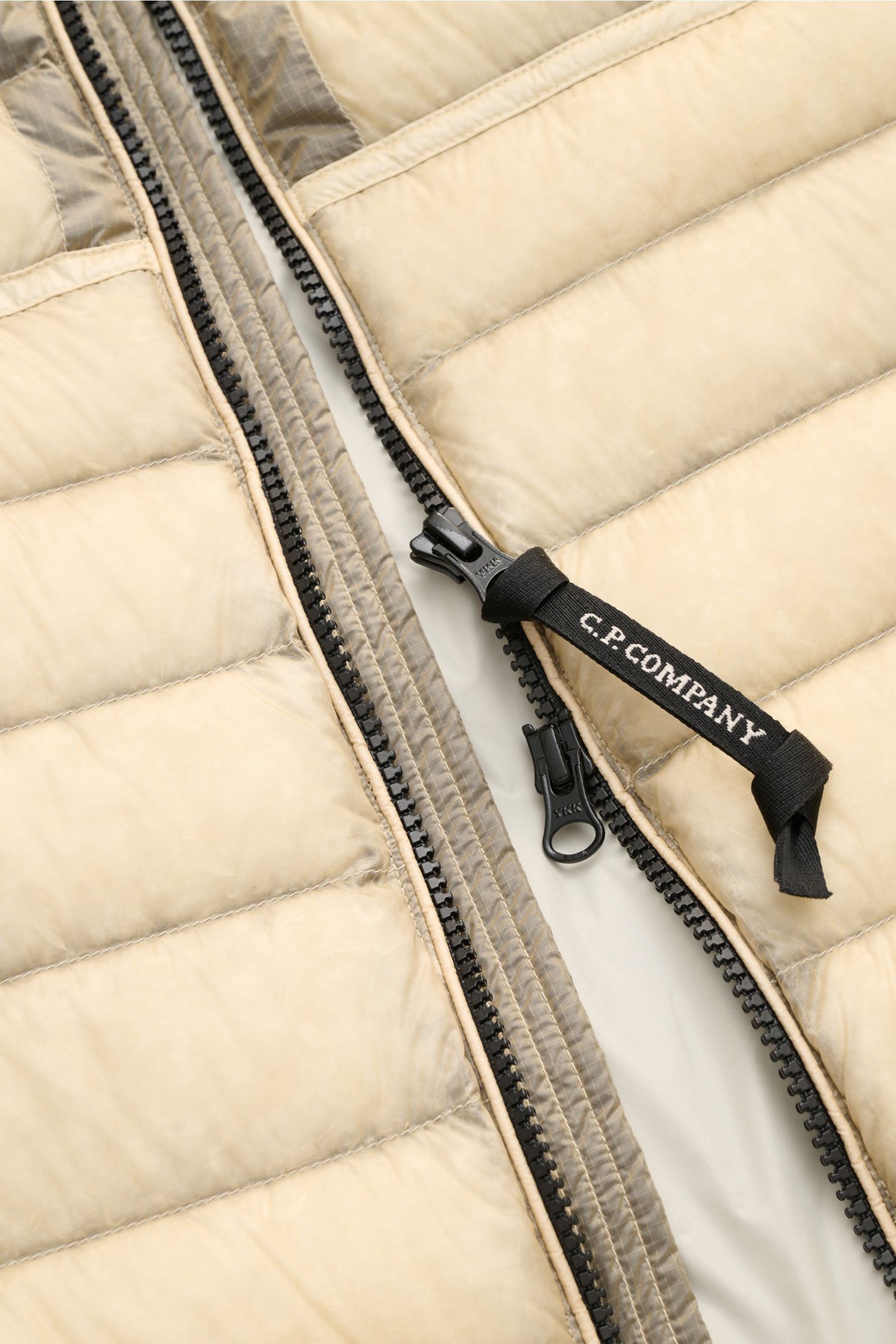 Close-up view of the zipper area of the C.P. Company Daunenweste 'D.D. Shell' beige, showing the black two-way zipper with branded pull tab on the superfine, water-repellent ripstop microfiber. The vest features a smooth touch, light down filling, windpro