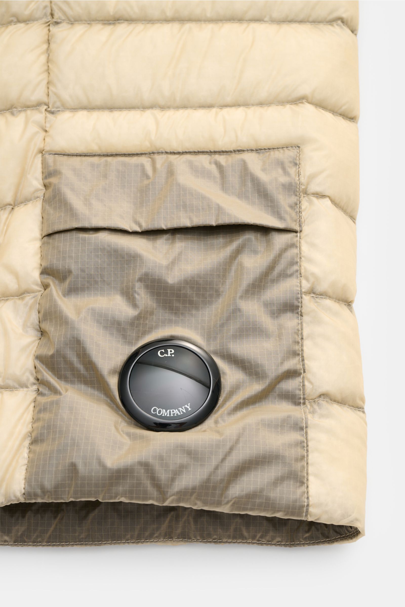 Close-up from the front of the beige C.P. Company Daunenweste 'D.D. Shell' showing a pocket with the signature lens detail on superfine, water-repellent ripstop-microfiber. The construction features lightweight down filling, windproof and garment-dyed fab