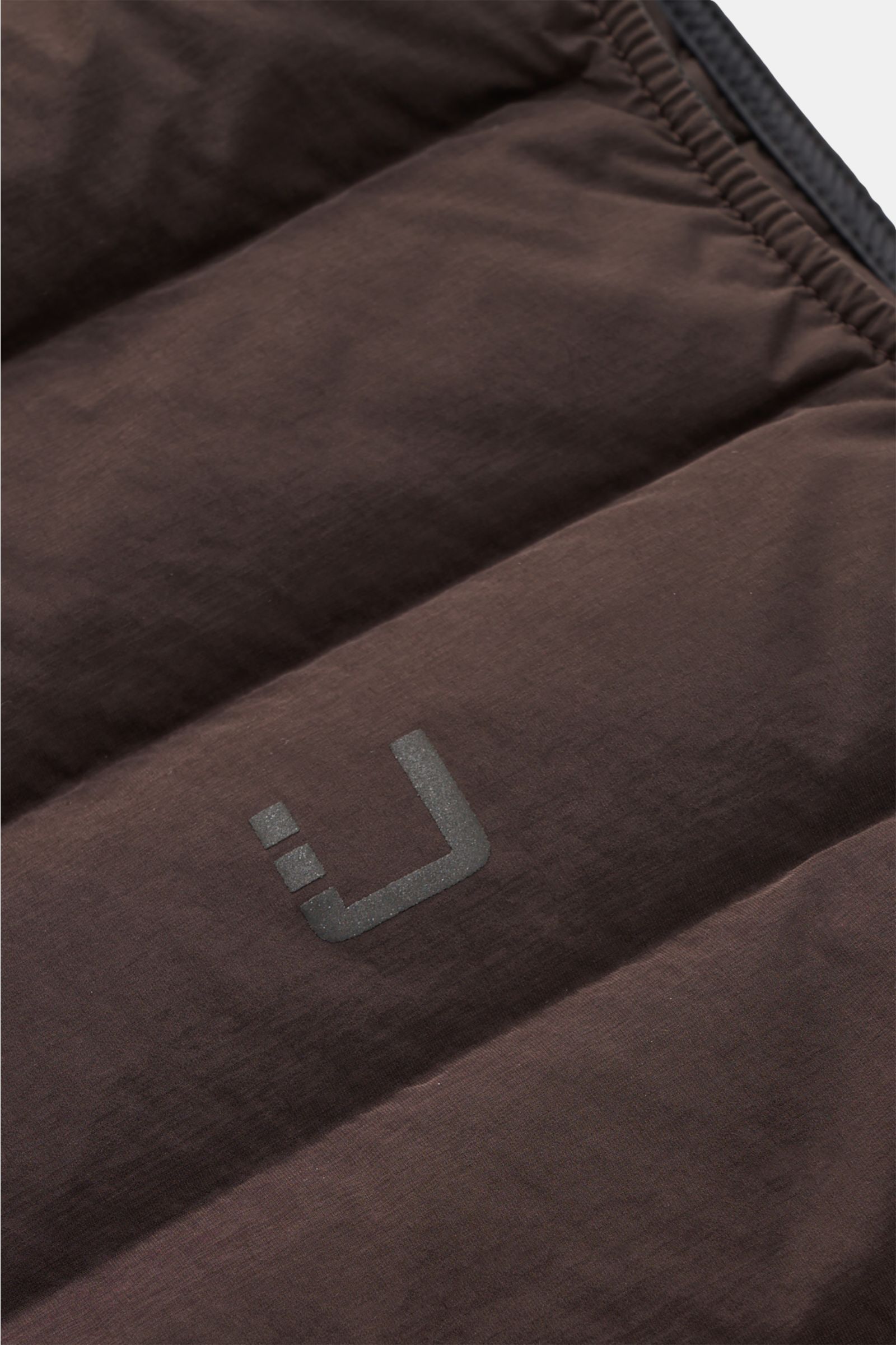 Close-up top-down view of the UBR Down gilet 'Supersonic' grey-brown showing the soft, stretchy microfibre with reflective logo print on the quilted, water-repellent fabric.