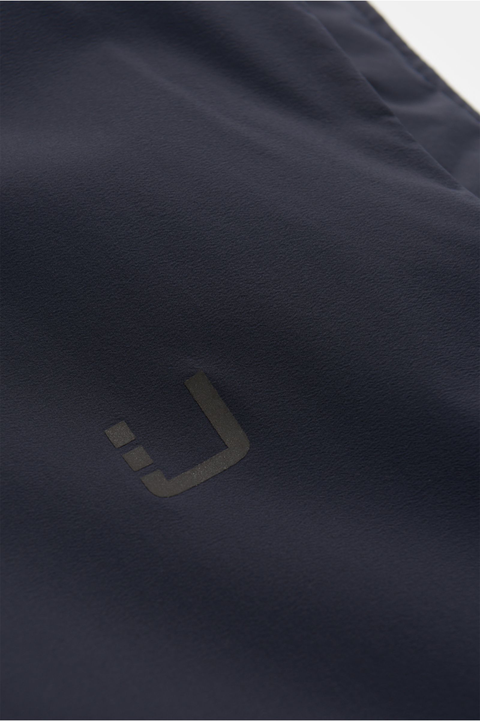 Close-up front view of UBR Outdoor-Weste 'Raptor' navy showing soft microfiber fabric with reflective logo print, lightweight padding, and smooth texture. Leichtigkeit, die man spürt – Performance, die bleibt: Mit der Weste 'Raptor' zeigt UBR, wie moderne