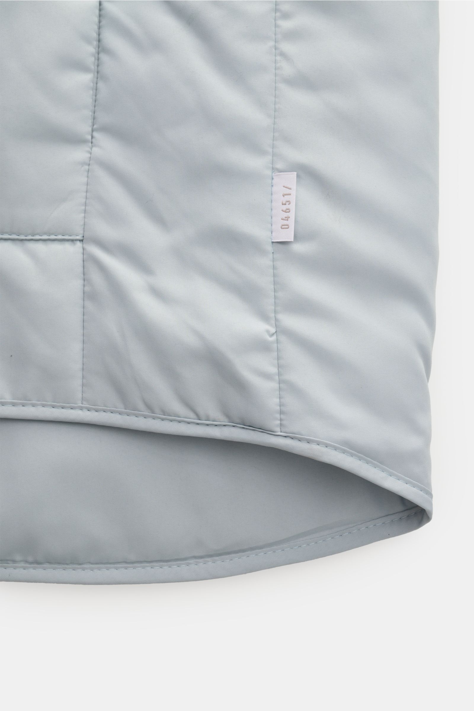 Close-up of the bottom hem and label of the 04651/ A trip in a bag Outdoor Weste 'MC26' hellgrau, showing light grey microfiber fabric with smooth texture and light padding, photographed from a front angle. Inspiriert von den Olympischen Winterspielen 