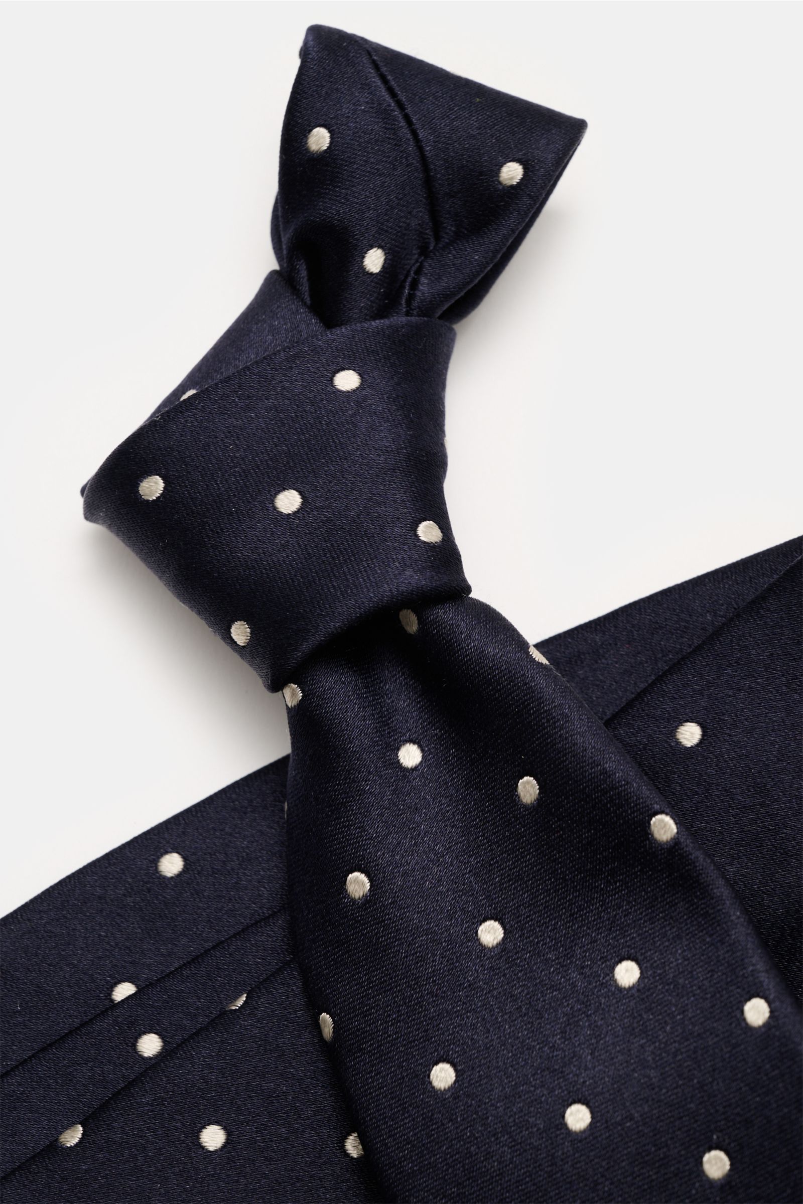 Close-up top view of a navy silk tie with cream polka dots, the Tom Ford Seidenkrawatte navy/creme gepunktet, showing its knot and fabric texture.

Zeitloser Stil mit luxuriöser Note: Die Seidenkrawatte von TOM FORD überzeugt als elegantes Essential fü