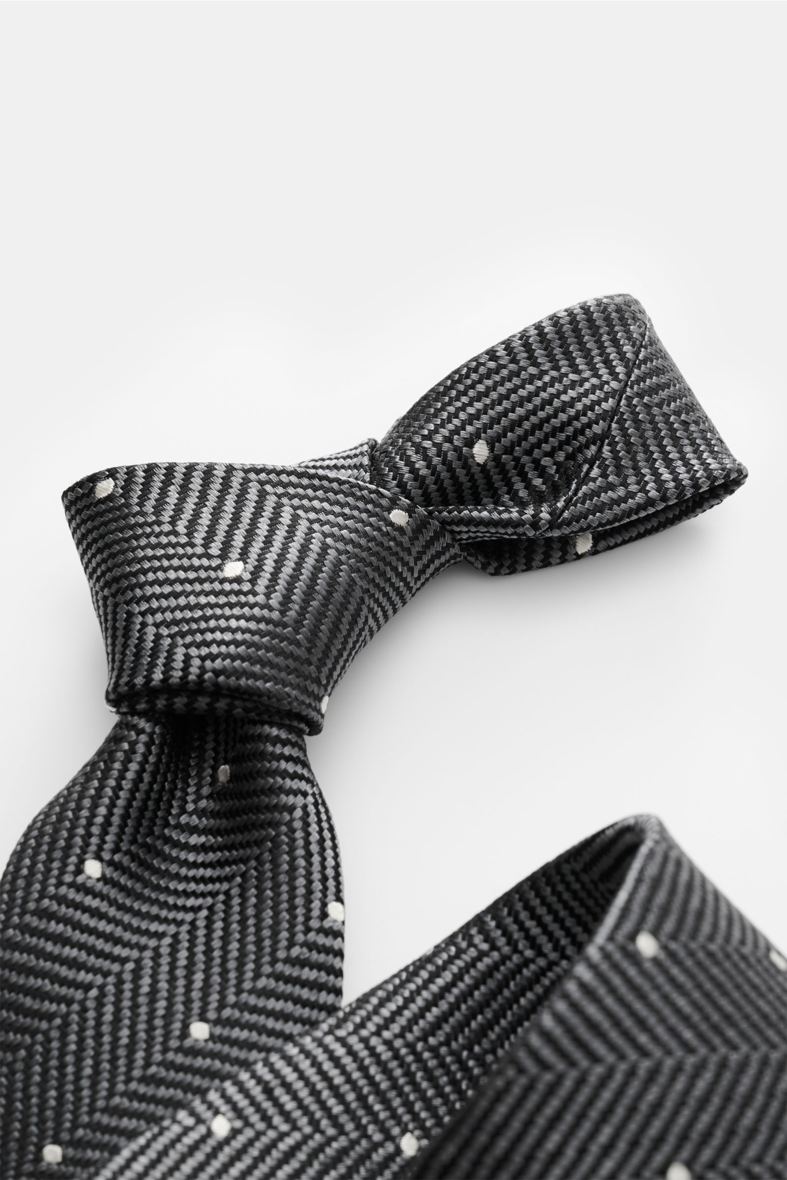 Tom Ford Seidenkrawatte dunkelgrau/schwarz/creme gemustert, in Detailaufnahme schräg von oben, Fischgratmuster mit Punkten, reine Seide, dezenter Glanz, 8 cm breit, made in Italy, Logo-Stitching.