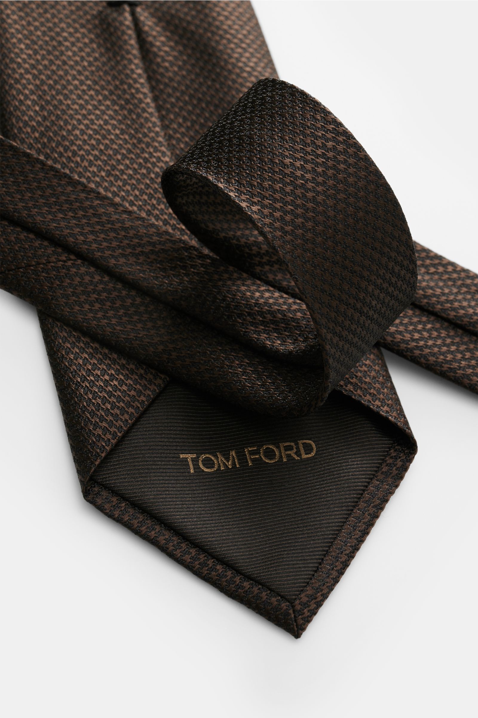 Tom Ford Seidenkrawatte dunkelbraun/schwarz gemustert mit zeitlosem Hahnentritt, aus reiner Seide, mit Logo-Stitching, Detailaufnahme von oben.