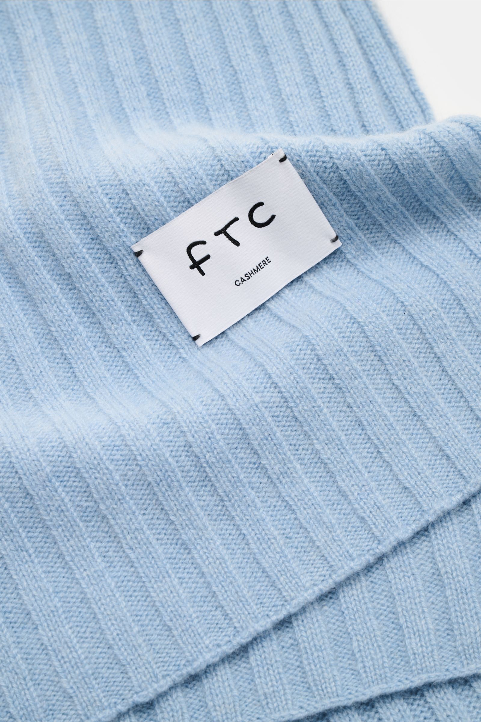 Nahaufnahme des FTC Cashmere Schals hellblau mit breitem Rippstrickmuster, großem Weblabel und weichem Griff aus reinem Fair Trade Cashmere.