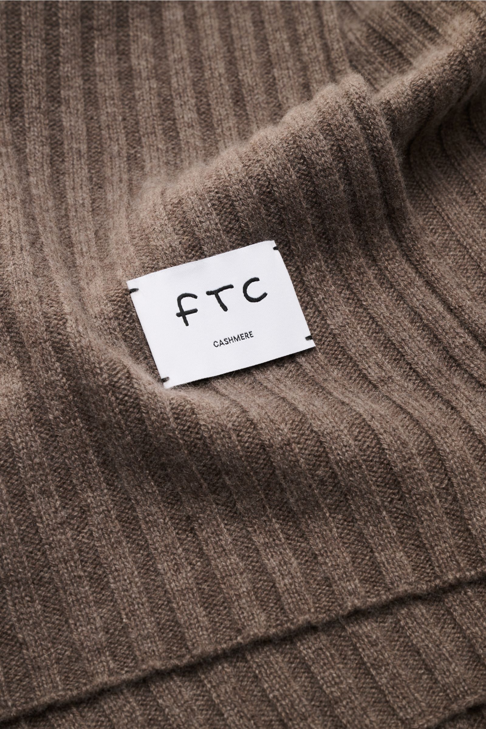 Nahaufnahme des graubraunen FTC Cashmere Schals mit breitem Rippstrickmuster und großem weißen Weblabel, weich und nachhaltig.