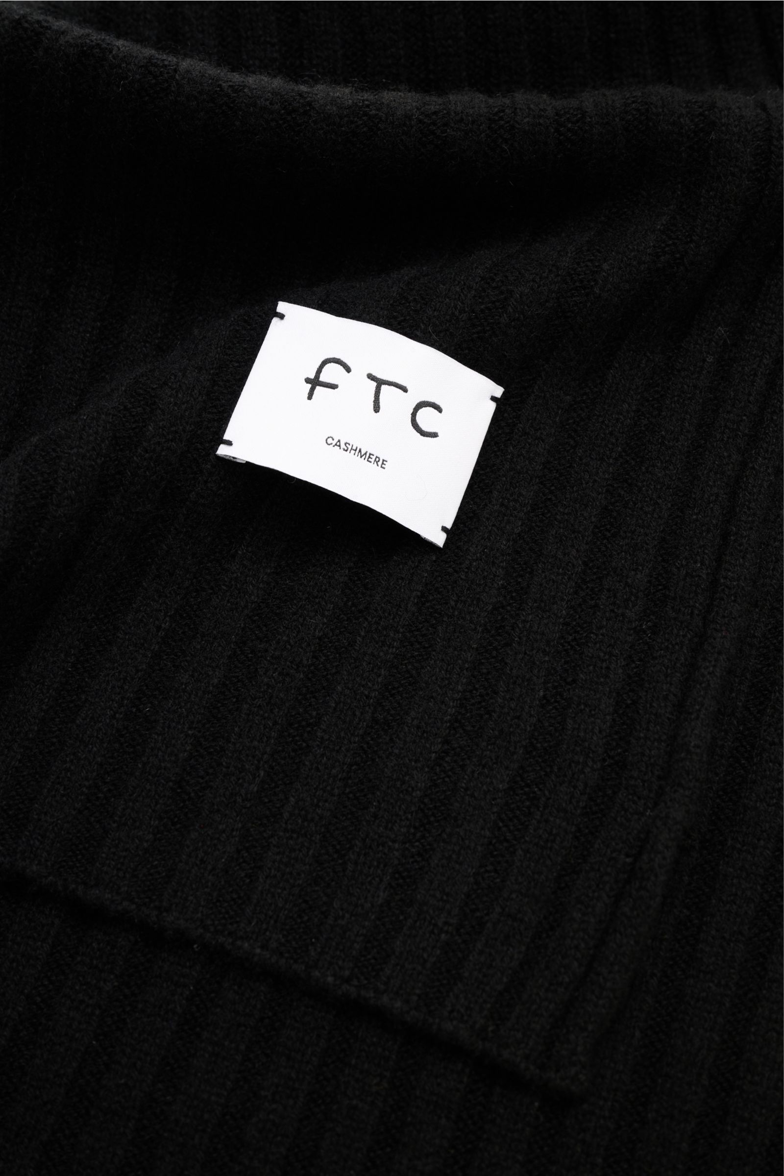 FTC Cashmere Schal schwarz in Nahaufnahme von oben fotografiert, breites Rippstrickmuster, weicher Griff, großes Weblabel mit Logo.